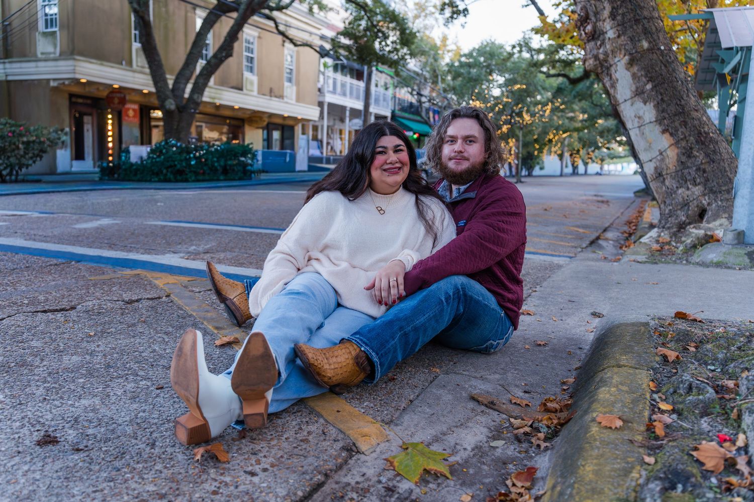 Two people sit together on fallen autumn leaves under a tree on a city sidewalk at sunset.
