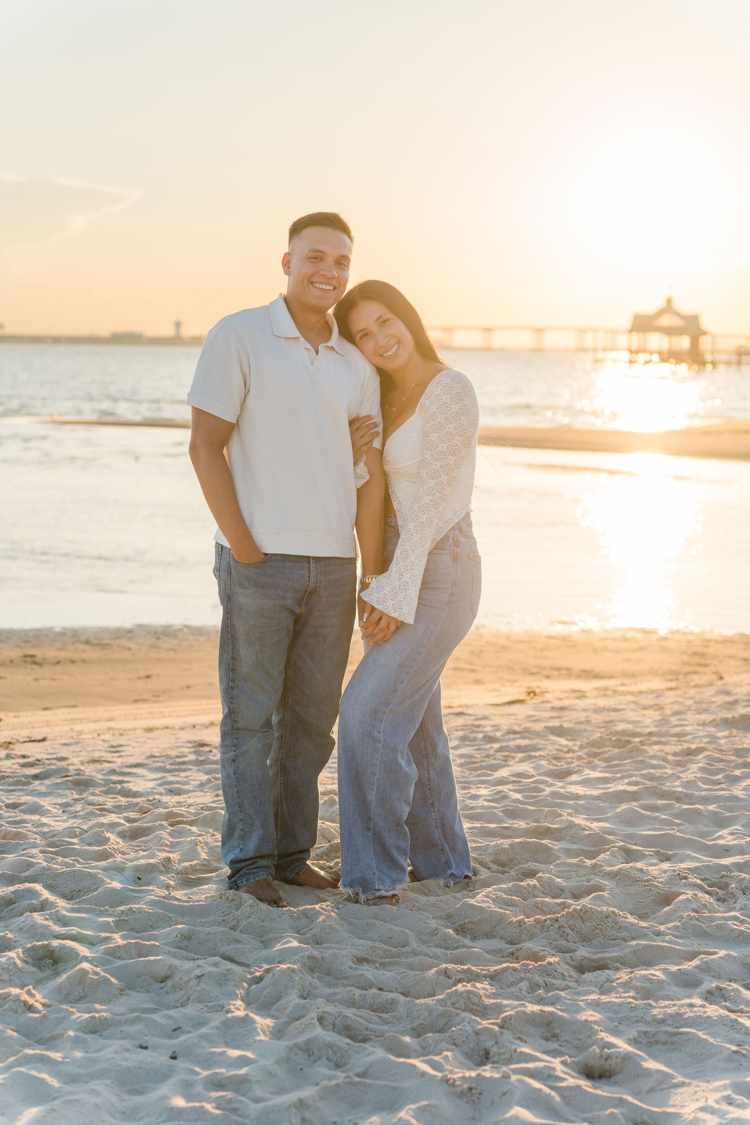 Romantic couple holds each other close on the beach with golden sunlight behind them.