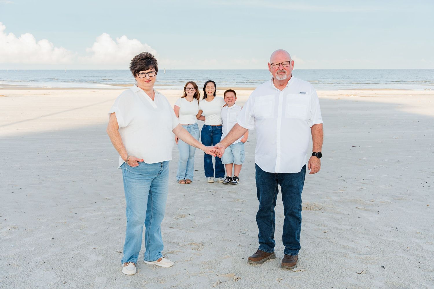 Family of five posing on a sandy beach, two adults in front and three children behind, with calm ocean waves in the background.