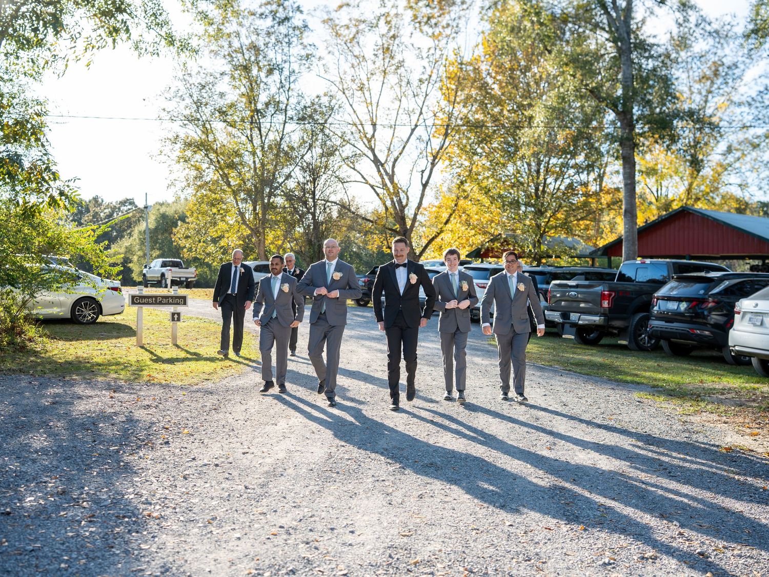 A group of people standing on a gravel path near parked cars and trees on a sunny autumn day.