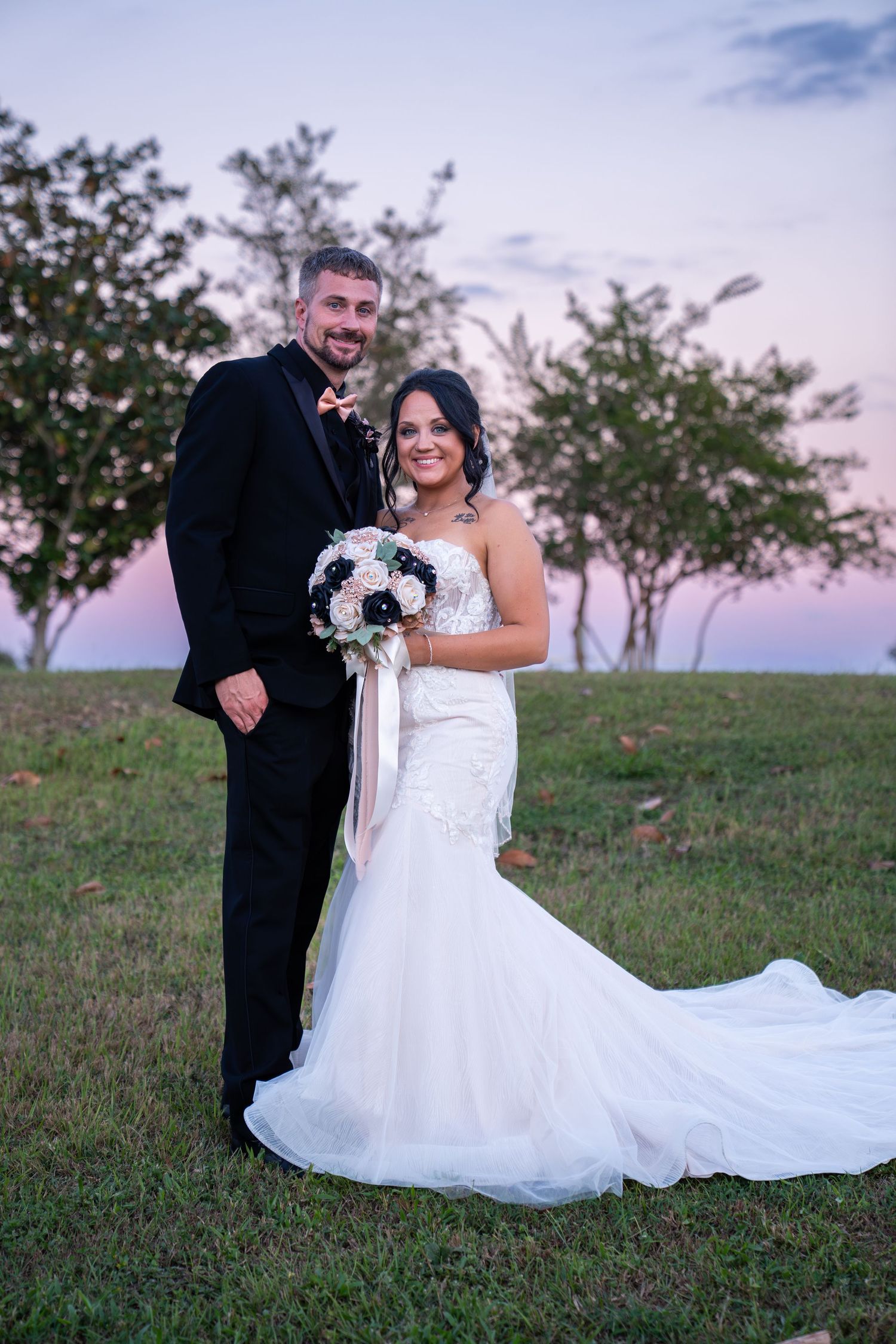 A romantic wedding photo of a couple in formal attire standing on a grassy hill at sunset with trees in the background.