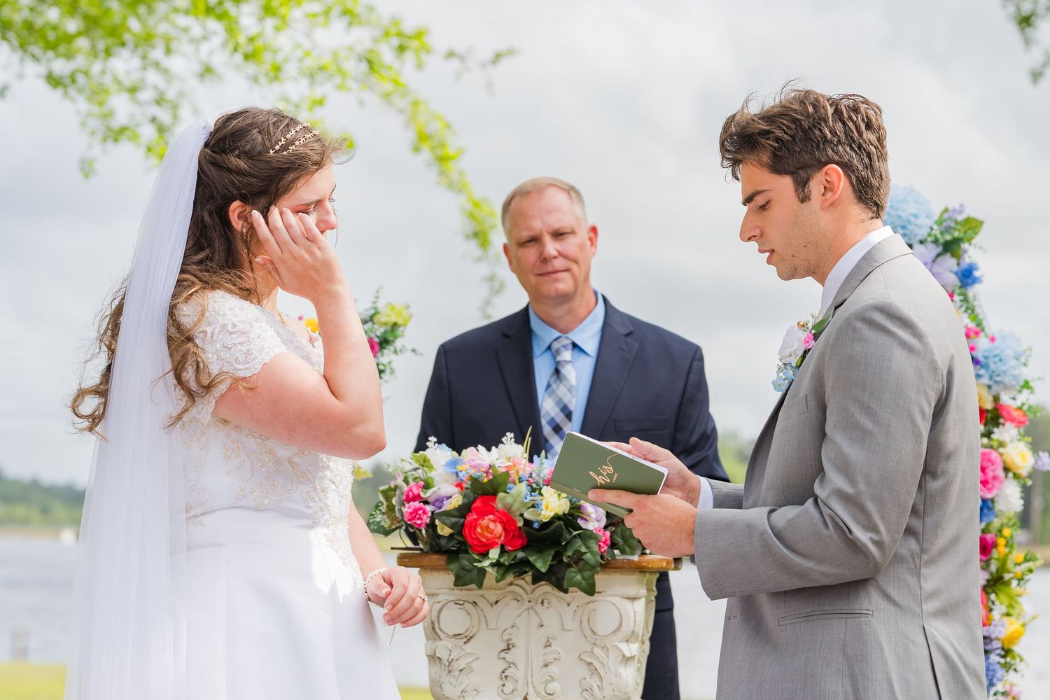 Emotional bride wiping tears as groom reads vows at outdoor wedding ceremony with officiant and floral arch.