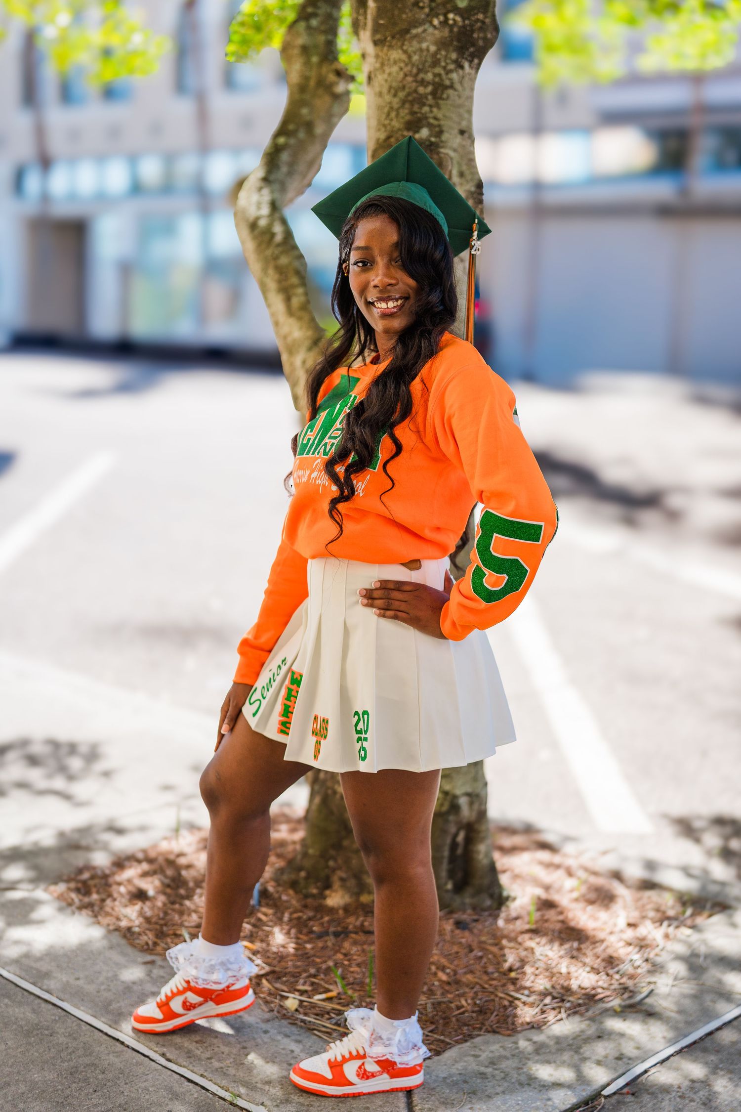 Graduate in bright orange jersey and white skirt smiles while posing outdoors.