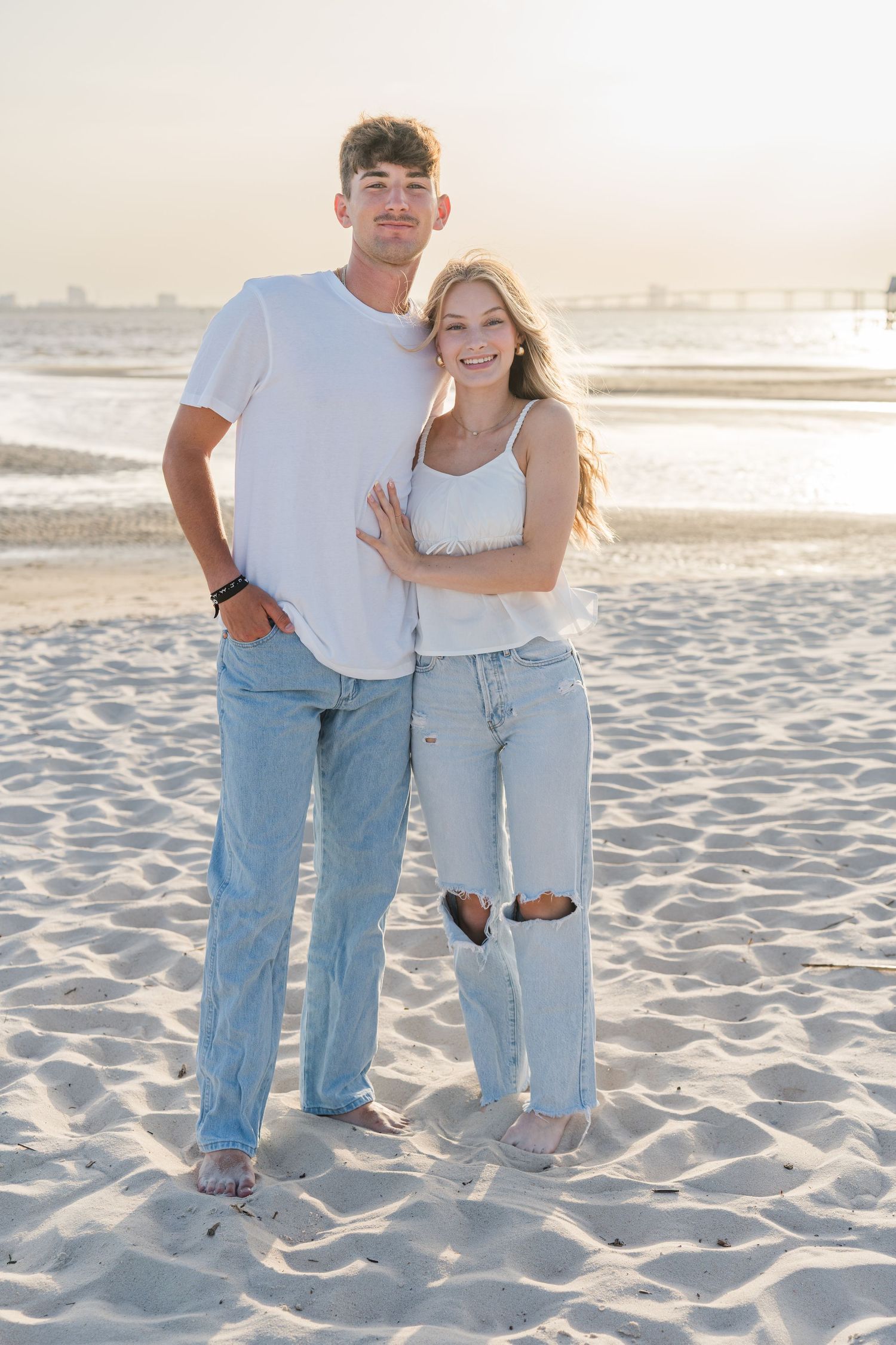 Couple in matching casual outfits poses together on sandy beach during sunset.