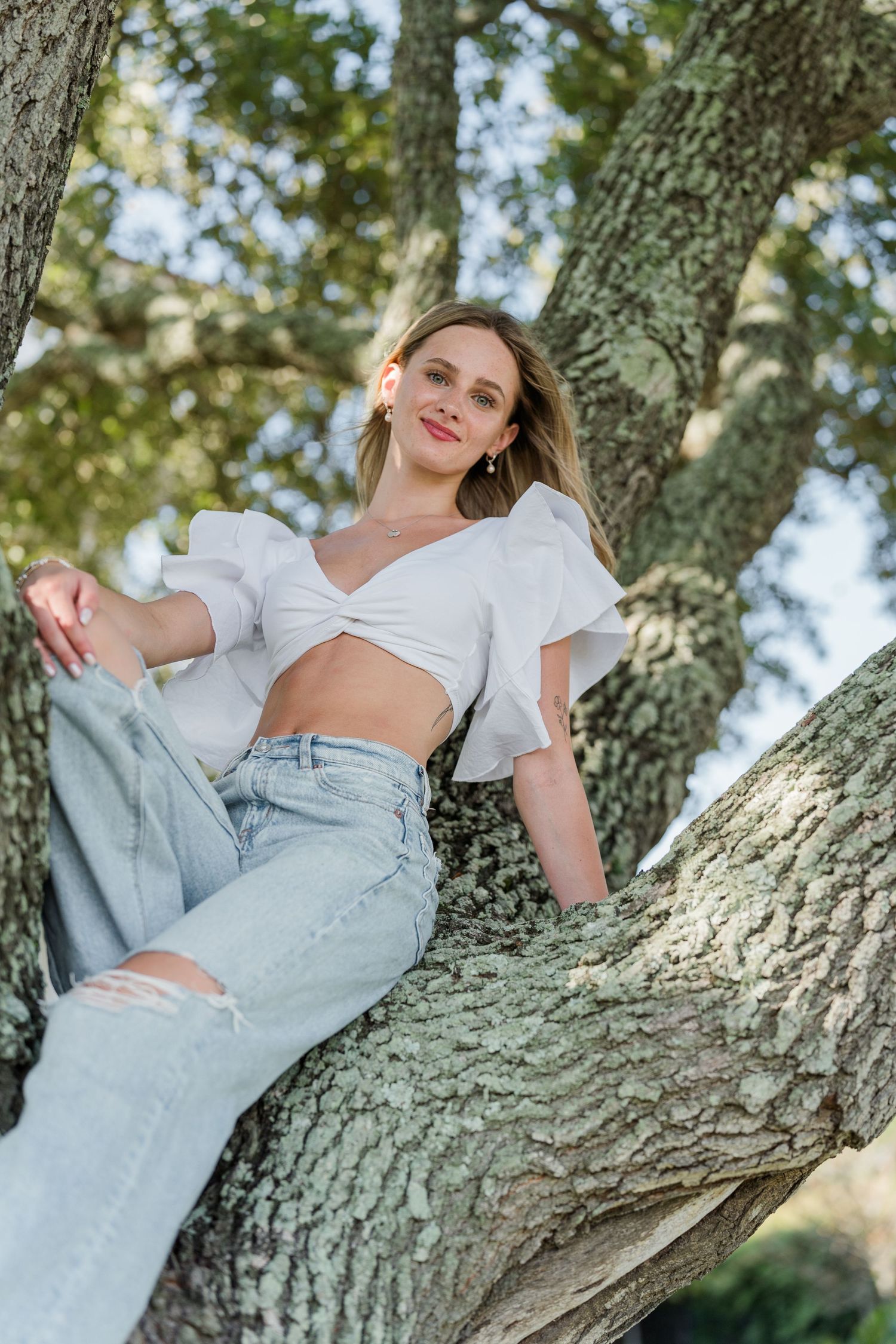 Person in white crop top and jeans reclines on tree branch in natural lighting.