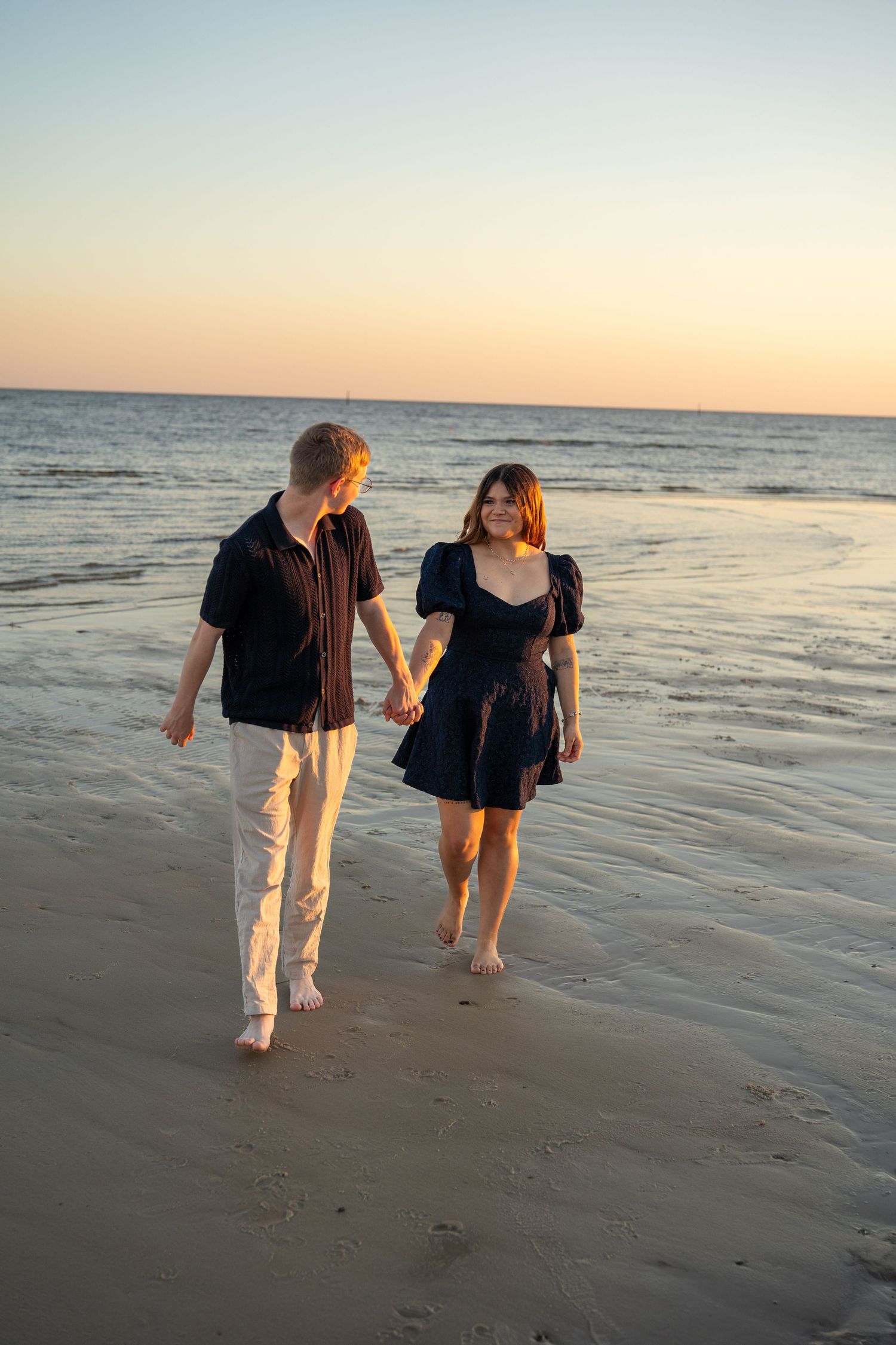 Two people walk hand in hand along the shoreline during golden hour at the beach.