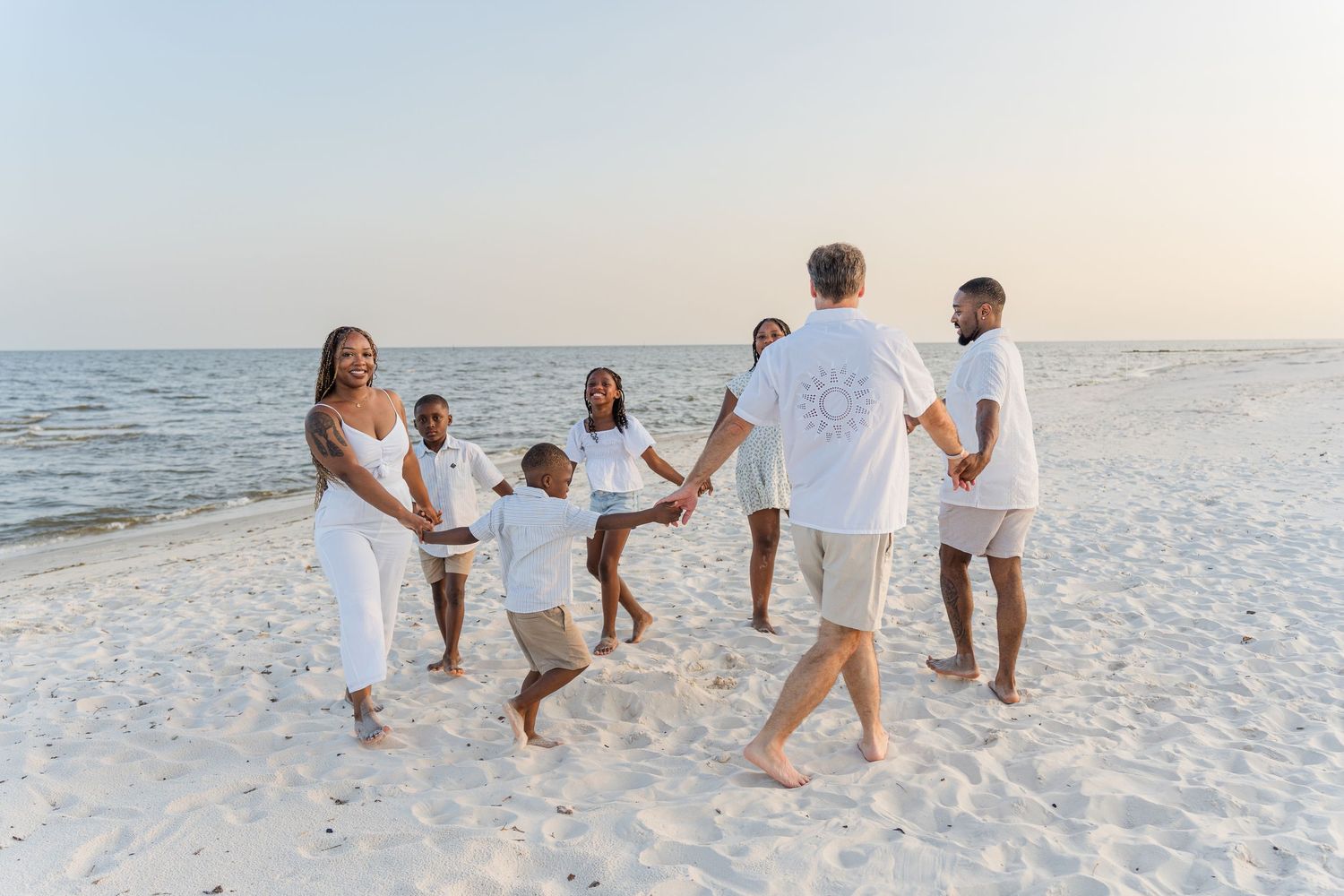 Extended family in white clothing walking together on a sandy beach at sunset.
