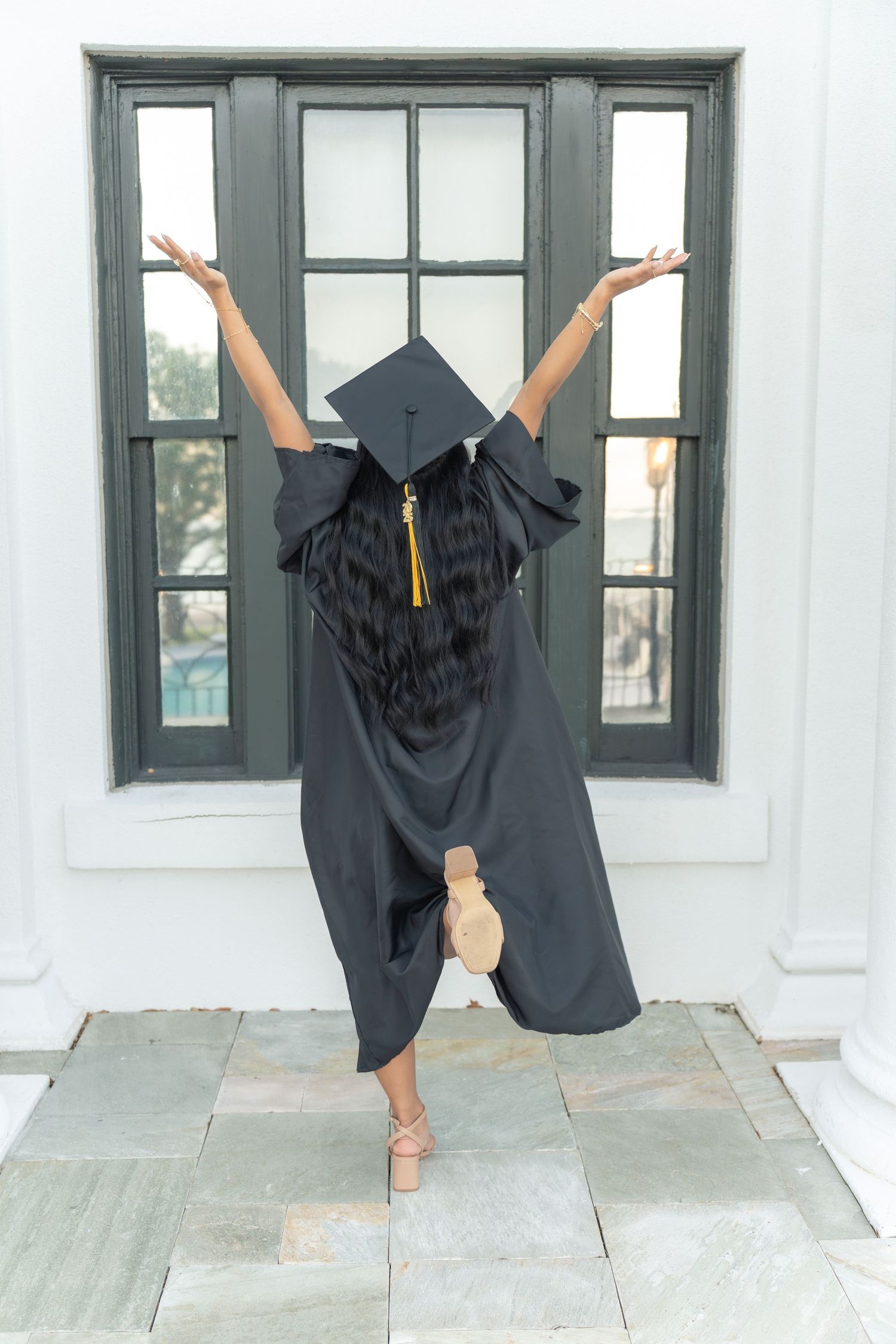 Fun graduation photoshoot showing black cap and gown against white wall and windows.