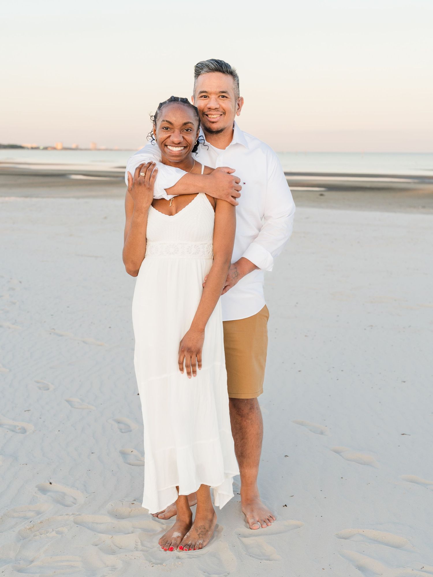A couple wearing white outfits poses together on a pristine beach at sunset.