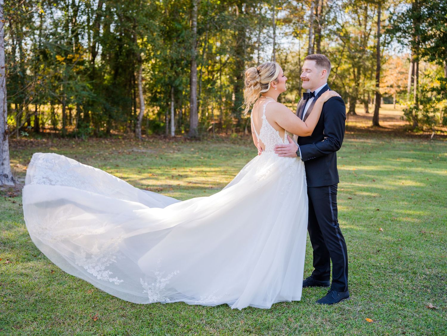 A couple in formal wedding attire embraces in a sunlit outdoor garden setting as the bride's veil flows gracefully in the breeze.