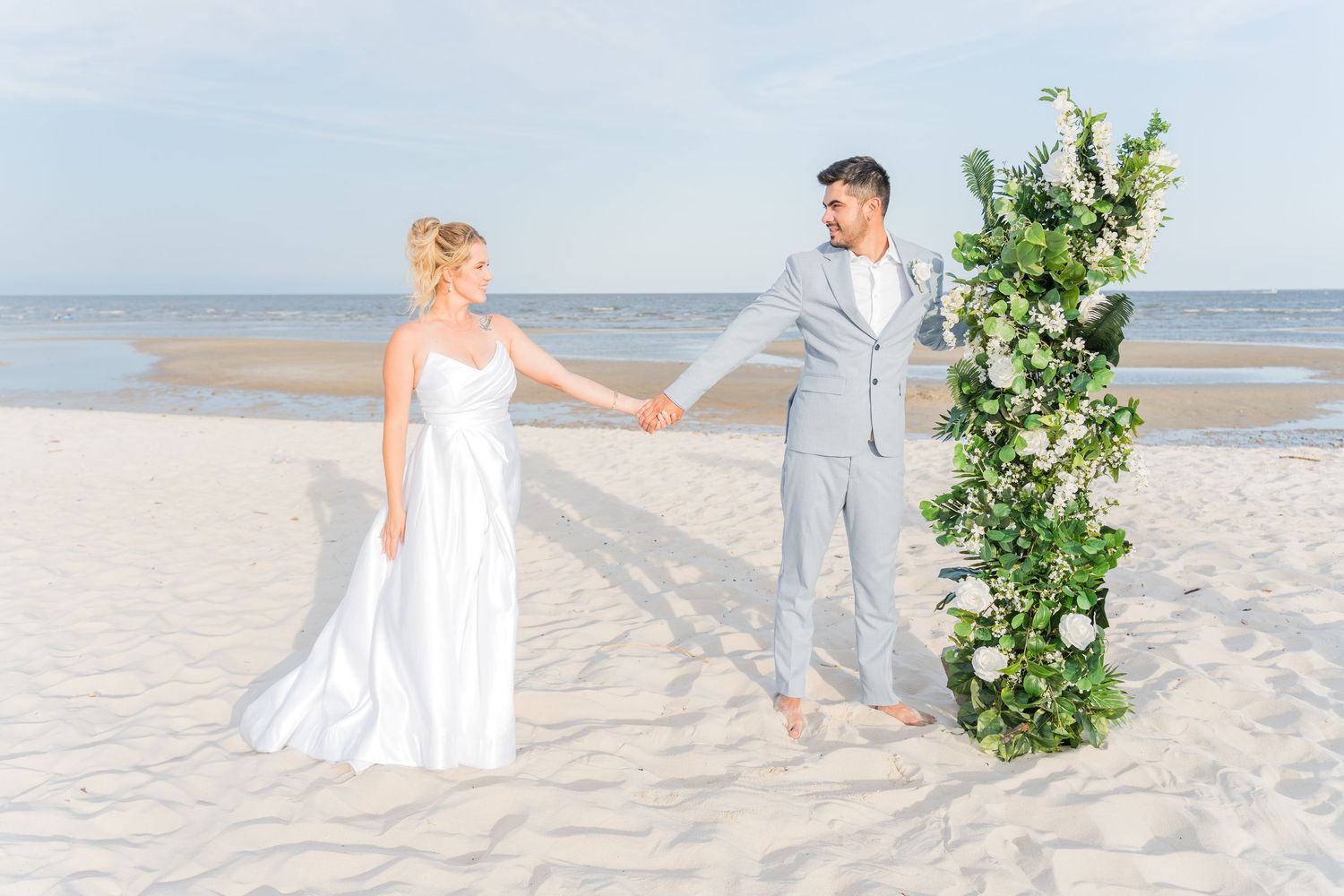Wedding couple walking hand in hand on a sandy beach with floral arrangements.