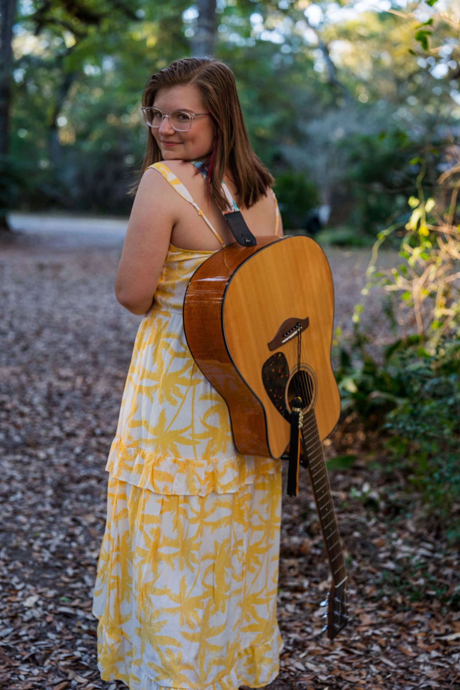 A person in a yellow dress holds an acoustic guitar while standing on a woodland path.