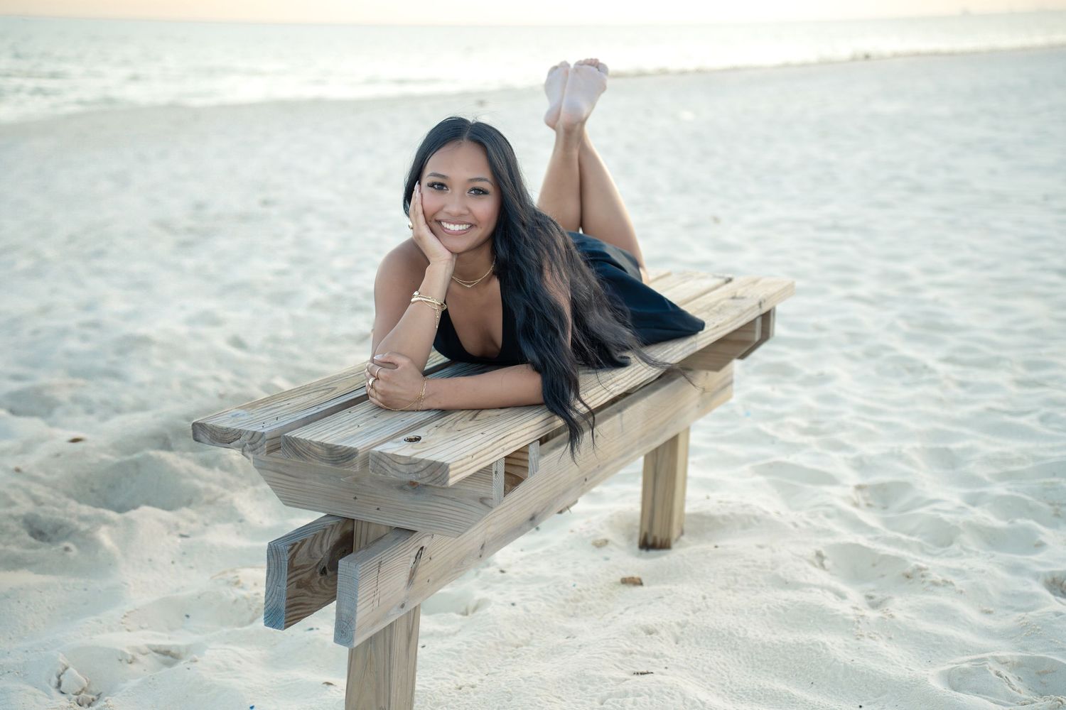 Person relaxing on wooden beach bench against sandy shoreline at sunset.