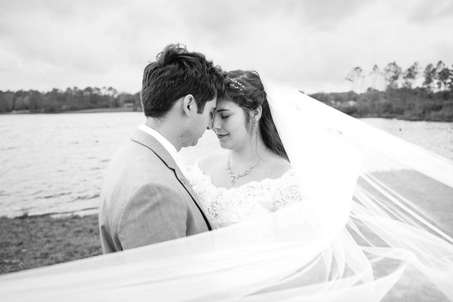 Black and white wedding photo of bride and groom nearly kissing by a lake, veil billowing in the wind.