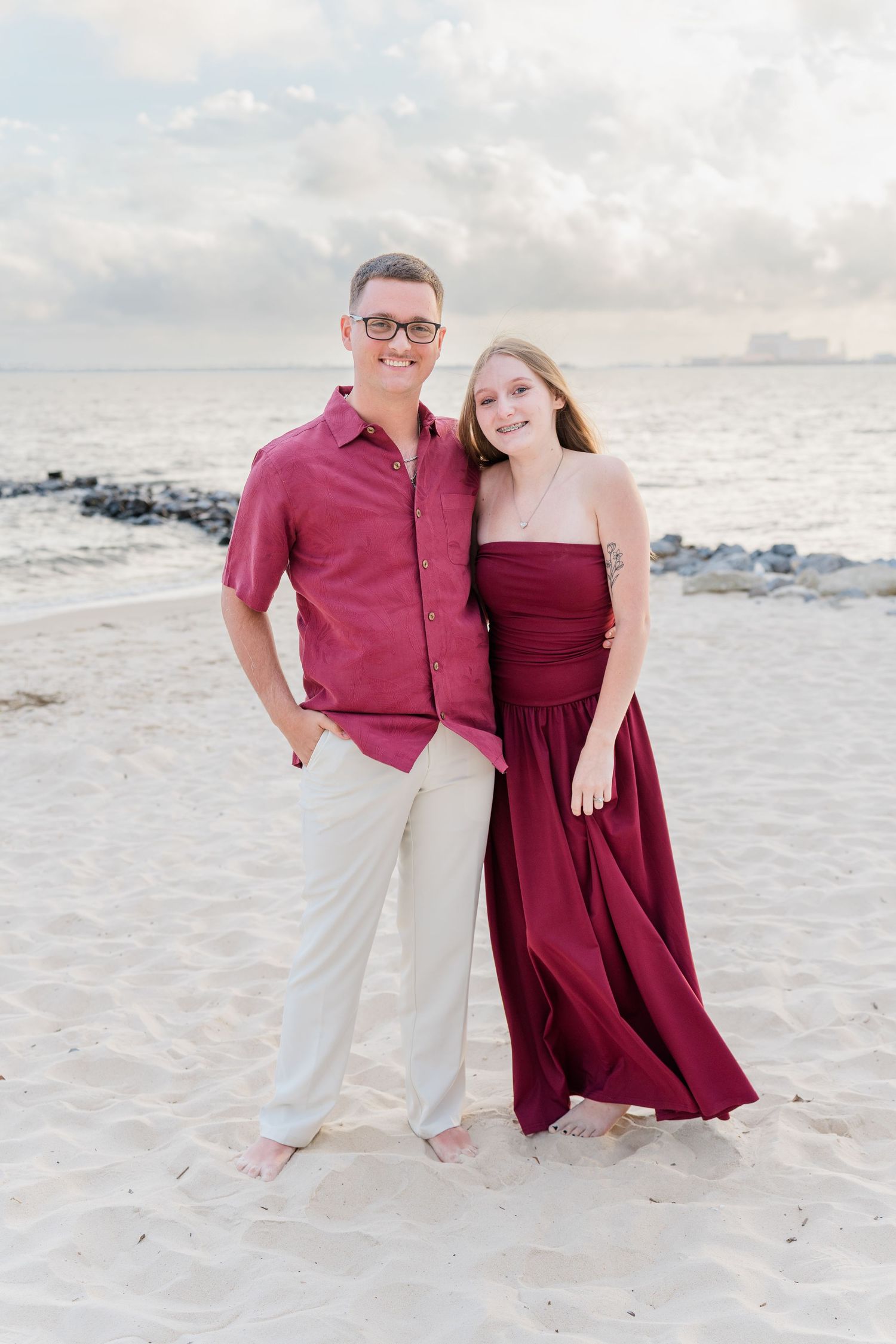 Couple in matching burgundy outfits posing on a sandy beach with ocean waves and cloudy sky backdrop.