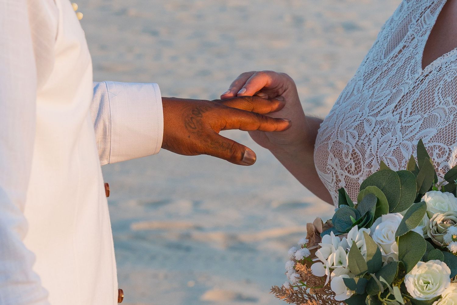 Close-up of hands exchanging rings during beach wedding ceremony with white roses.