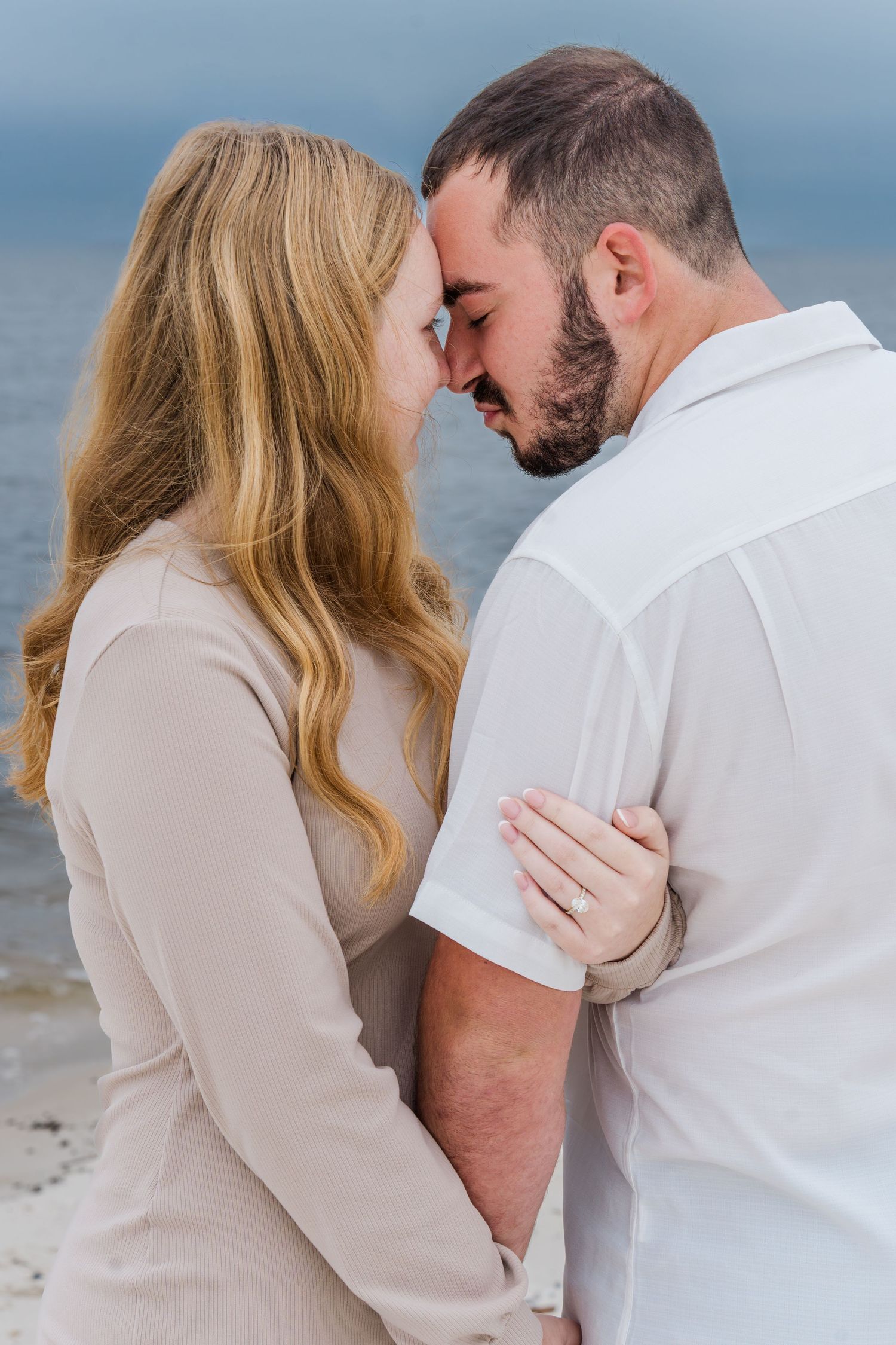 A romantic couple embraces on a sandy beach while wearing white and beige clothing against an ocean backdrop.