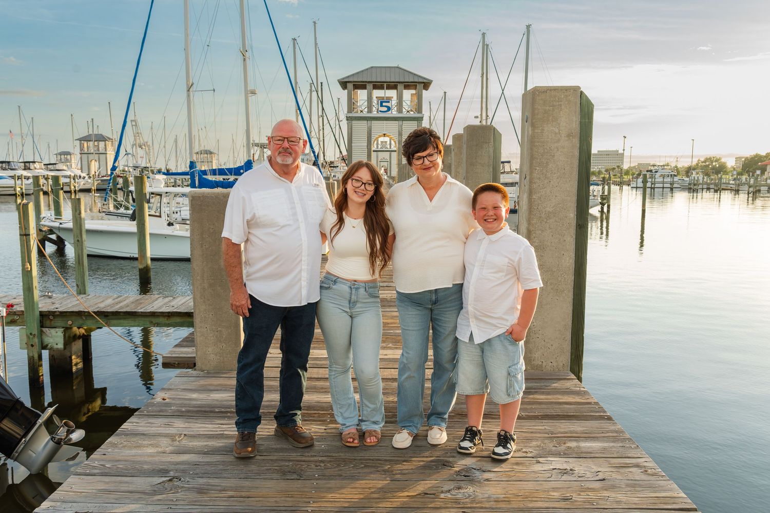 Family of four smiling on a marina dock with sailboats and calm water in the background on a sunny day.