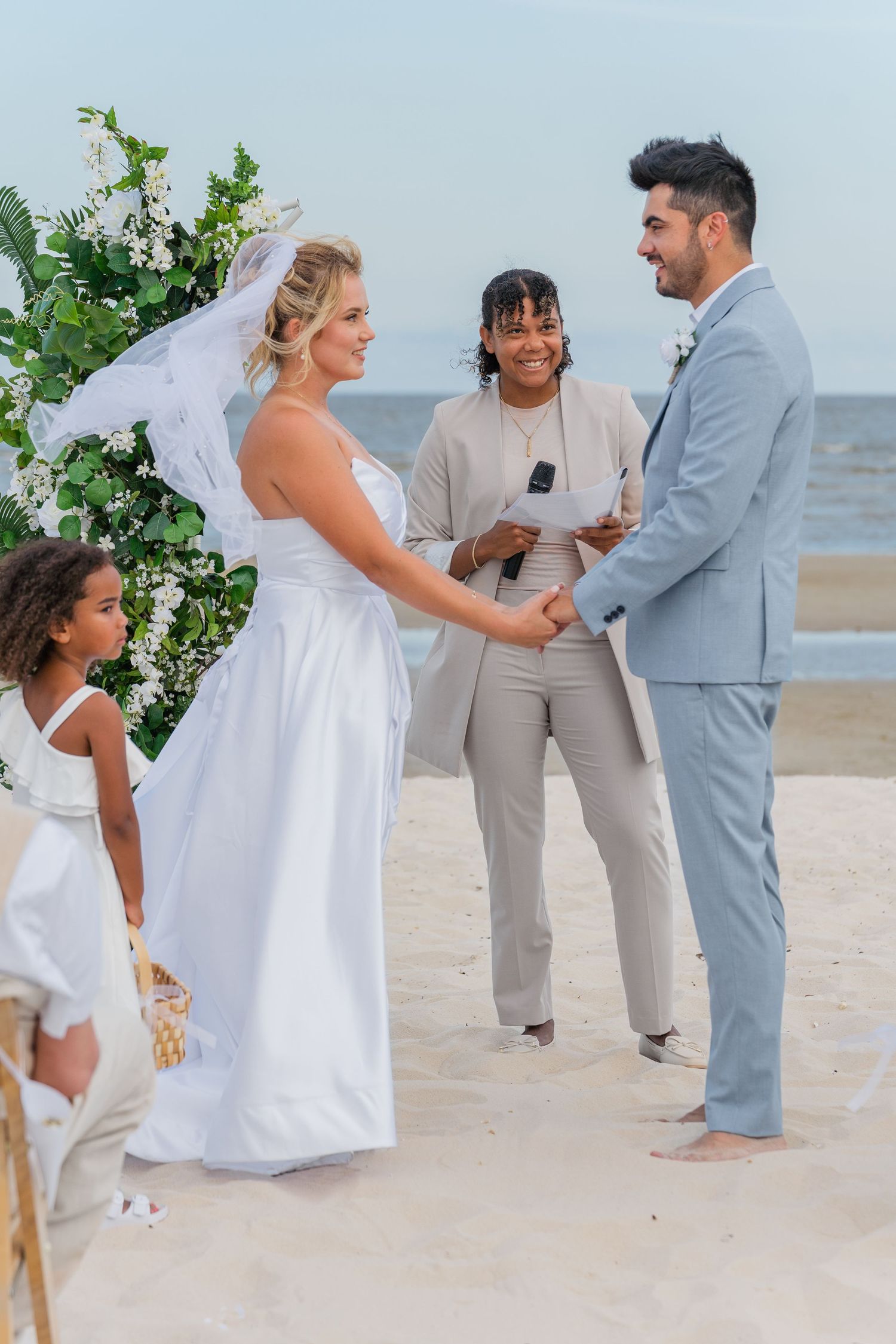 A beach wedding ceremony in progress with guests wearing white and grey attire.