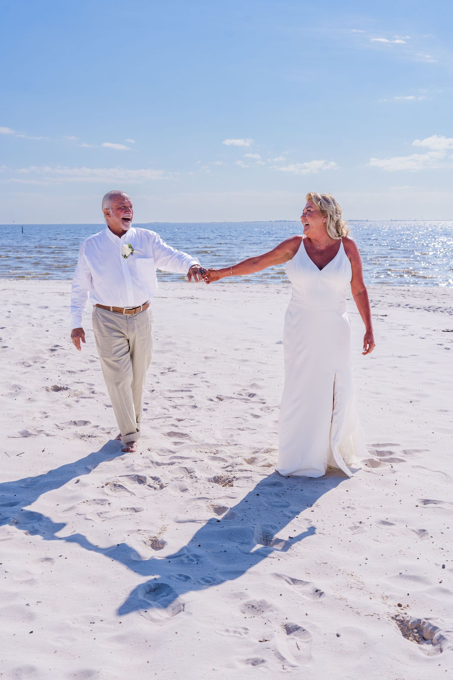 A couple in white attire dances together on a pristine beach with bright blue ocean and sky in the background.