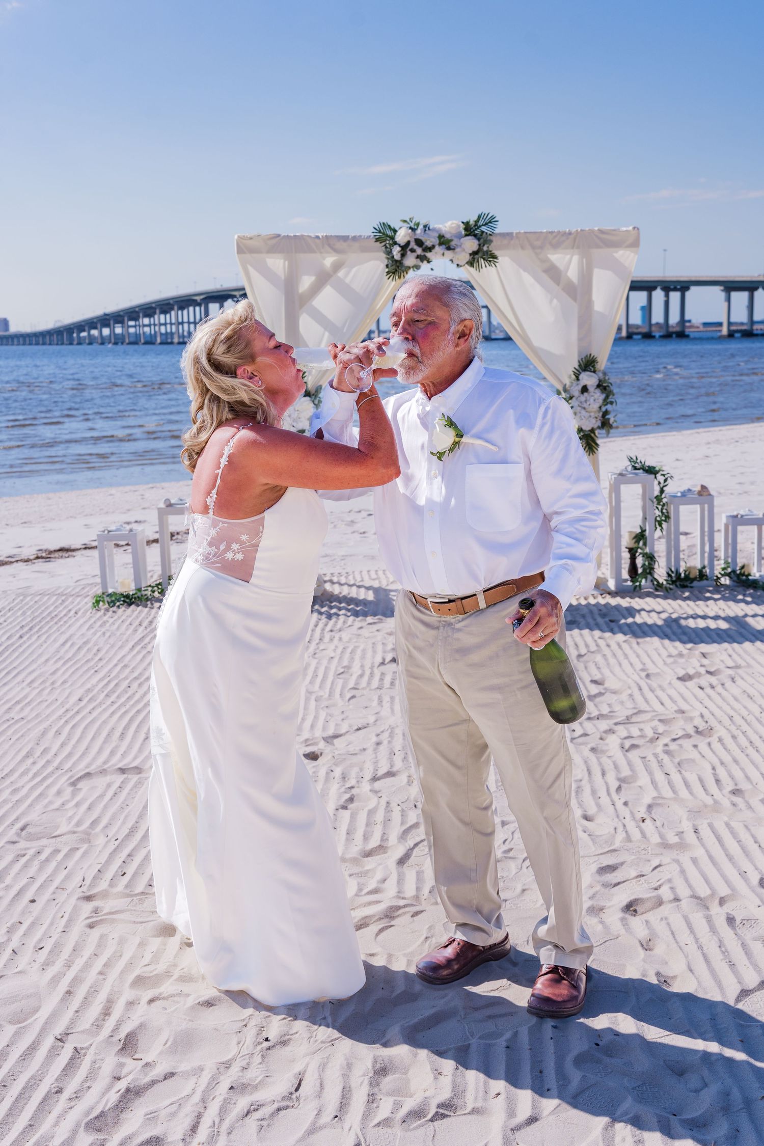 A couple shares a celebratory drink on a sunny beach during their wedding ceremony with a bridge visible in the background.