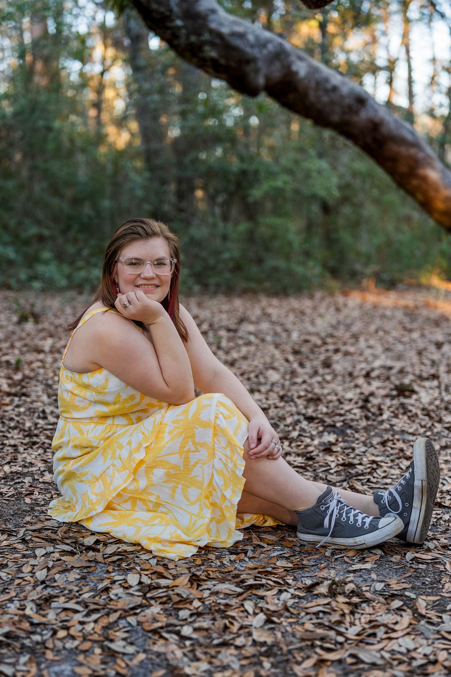A person in a yellow summer dress and sneakers sits on fallen leaves in a wooded setting.