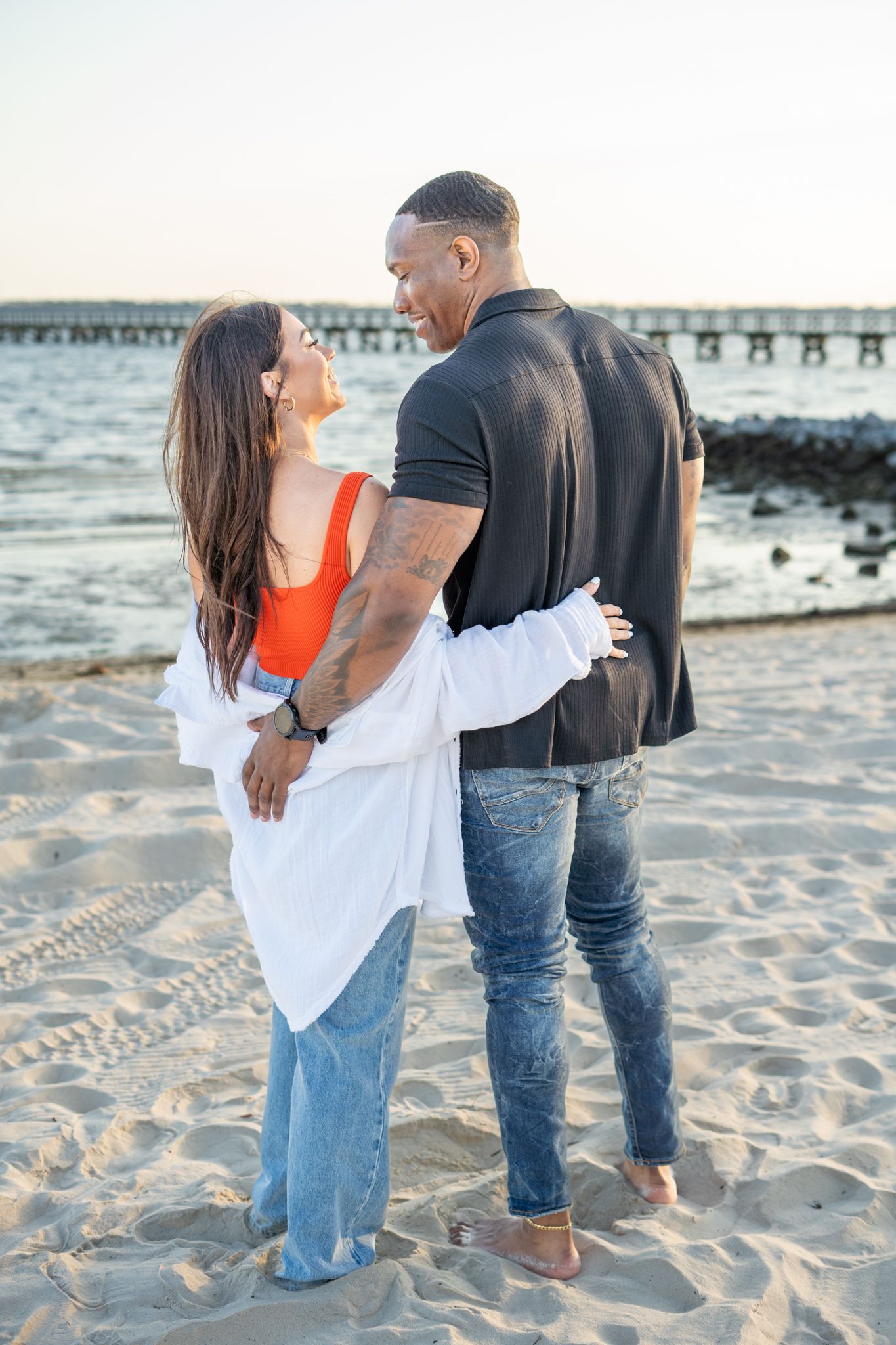 A couple shares an intimate moment on the beach with a pier and ocean waves in the background.
