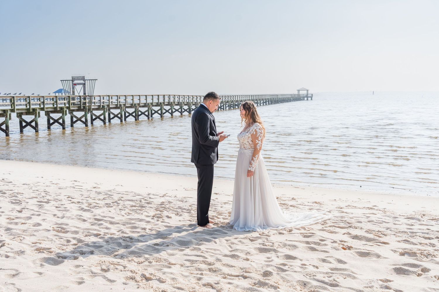 A couple stands together on a pristine white sandy beach with a wooden pier stretching into the ocean behind them.