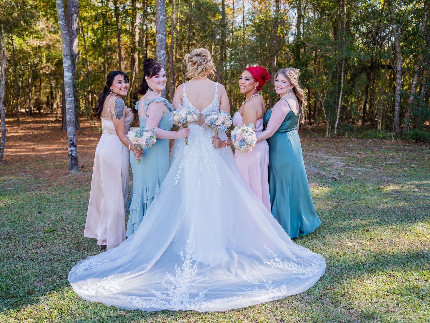 A bride and bridesmaids in pink and blue dresses gather in a sun-dappled forest setting during a wedding celebration.