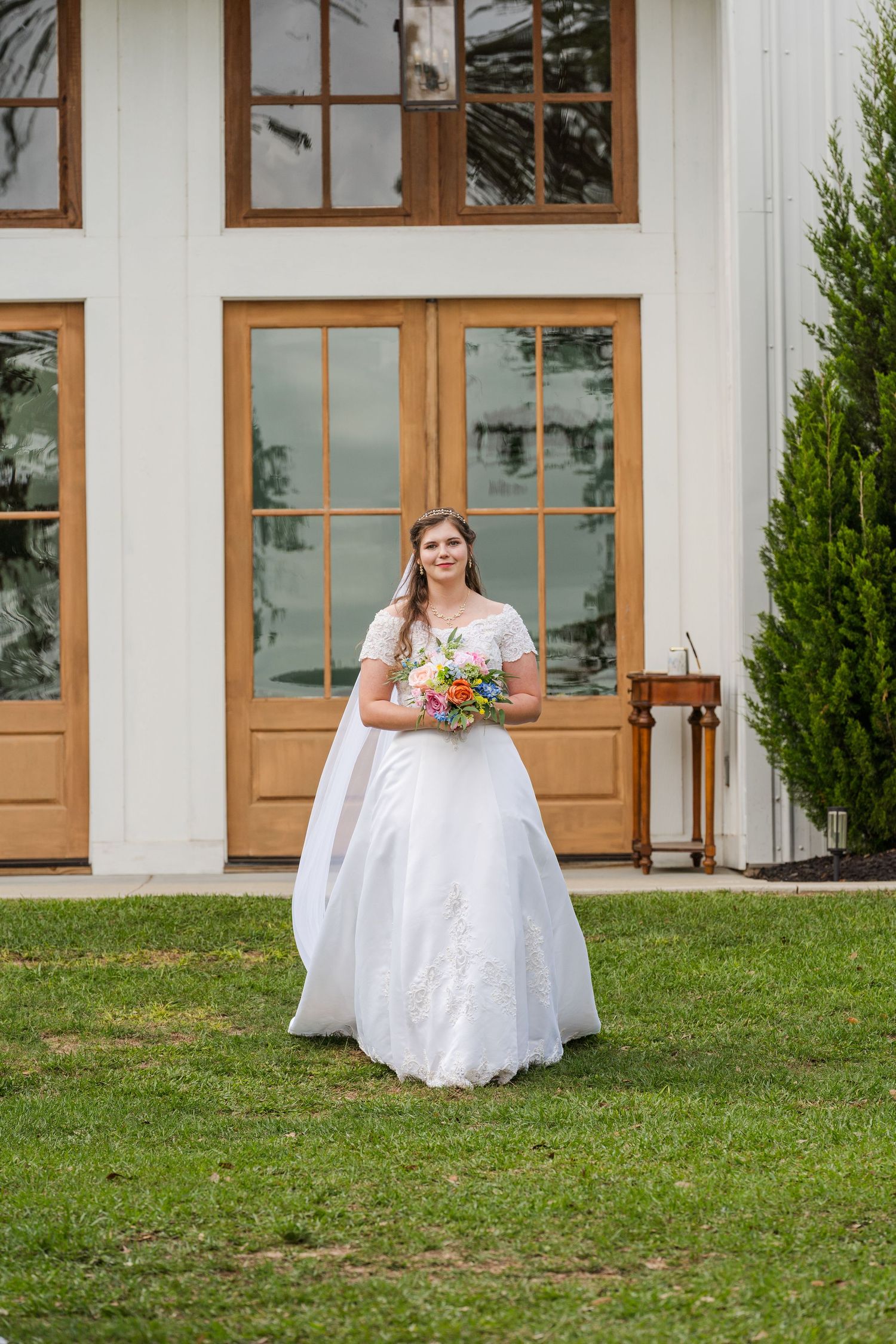 Bride in white gown holding colorful bouquet stands on green lawn before elegant white building with wooden French doors.