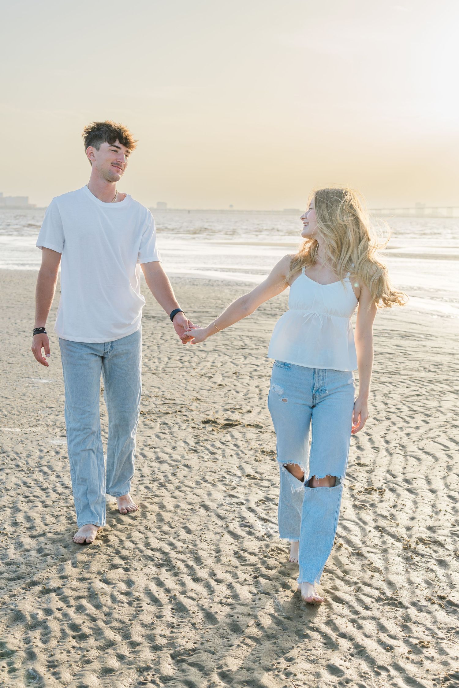 Couple walking hand in hand on the beach wearing white shirts and light denim.