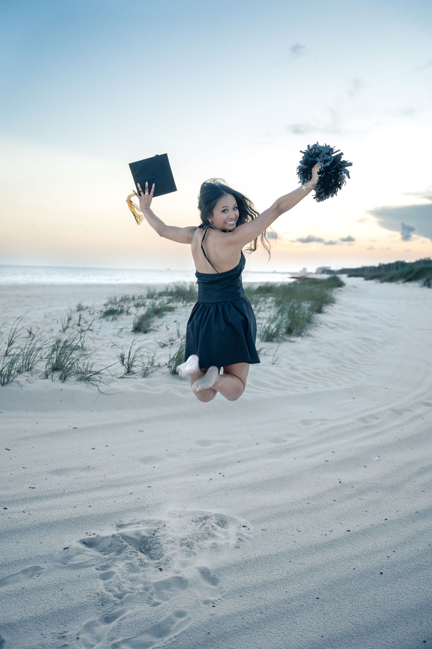 Graduate jumping and celebrating with cap on sandy beach at dusk.