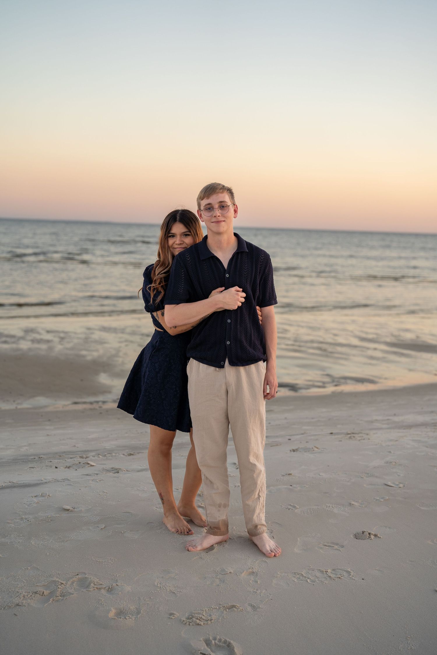 A couple stands together barefoot on a peaceful beach during sunset.