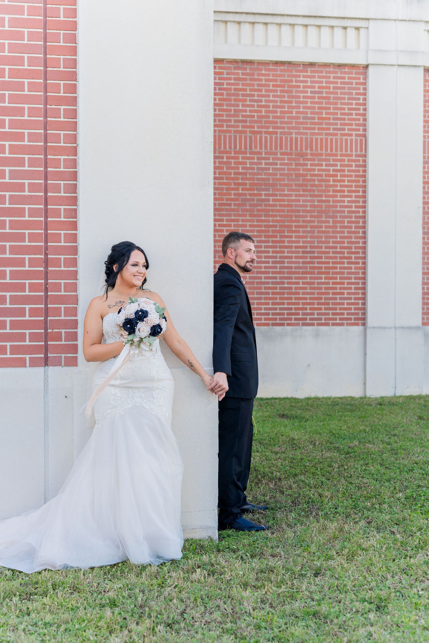 A wedding couple poses playfully on opposite sides of a brick wall while holding hands in a first look photo.