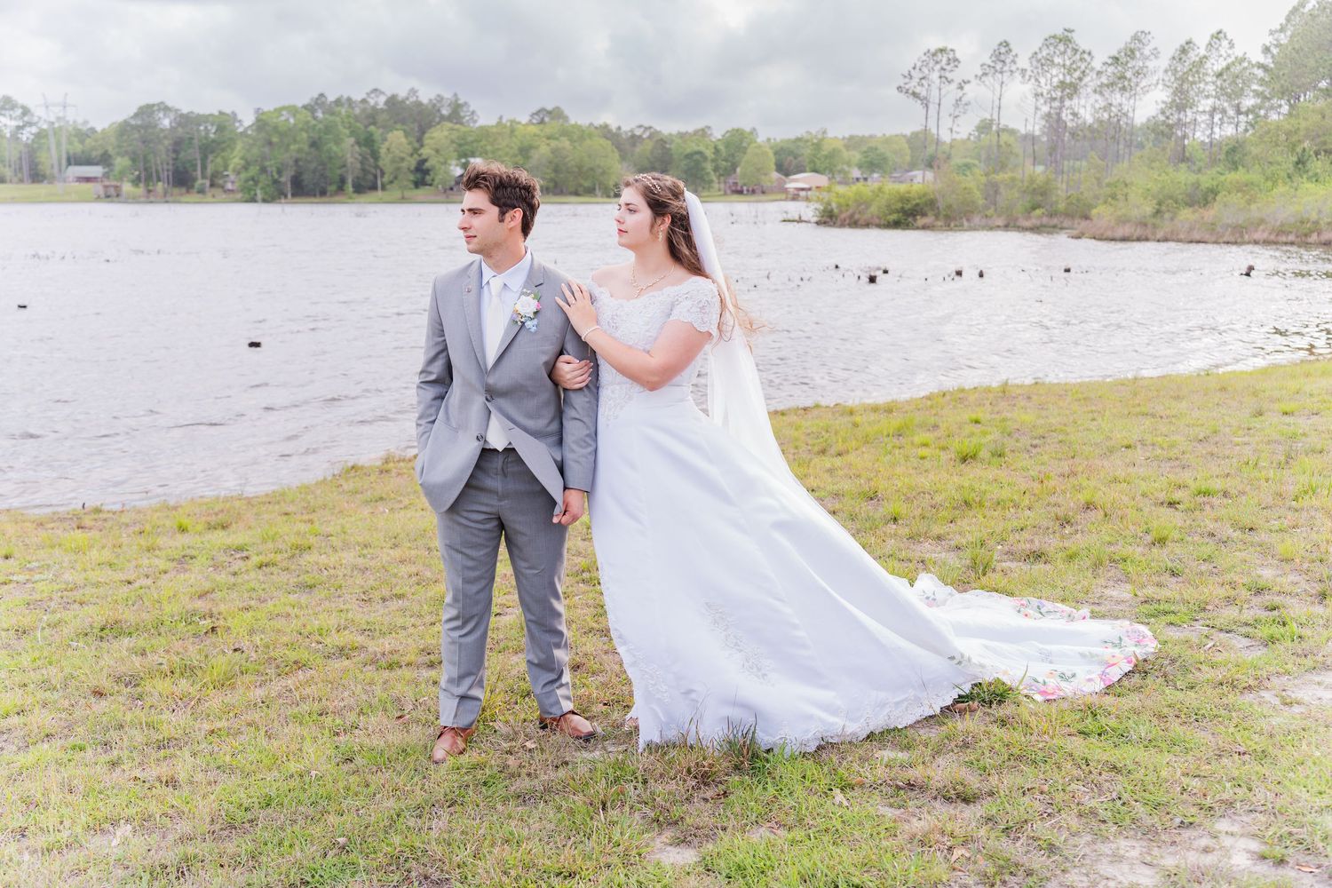 Bride in white gown and groom in gray suit hold hands by a lakeside on their wedding day.