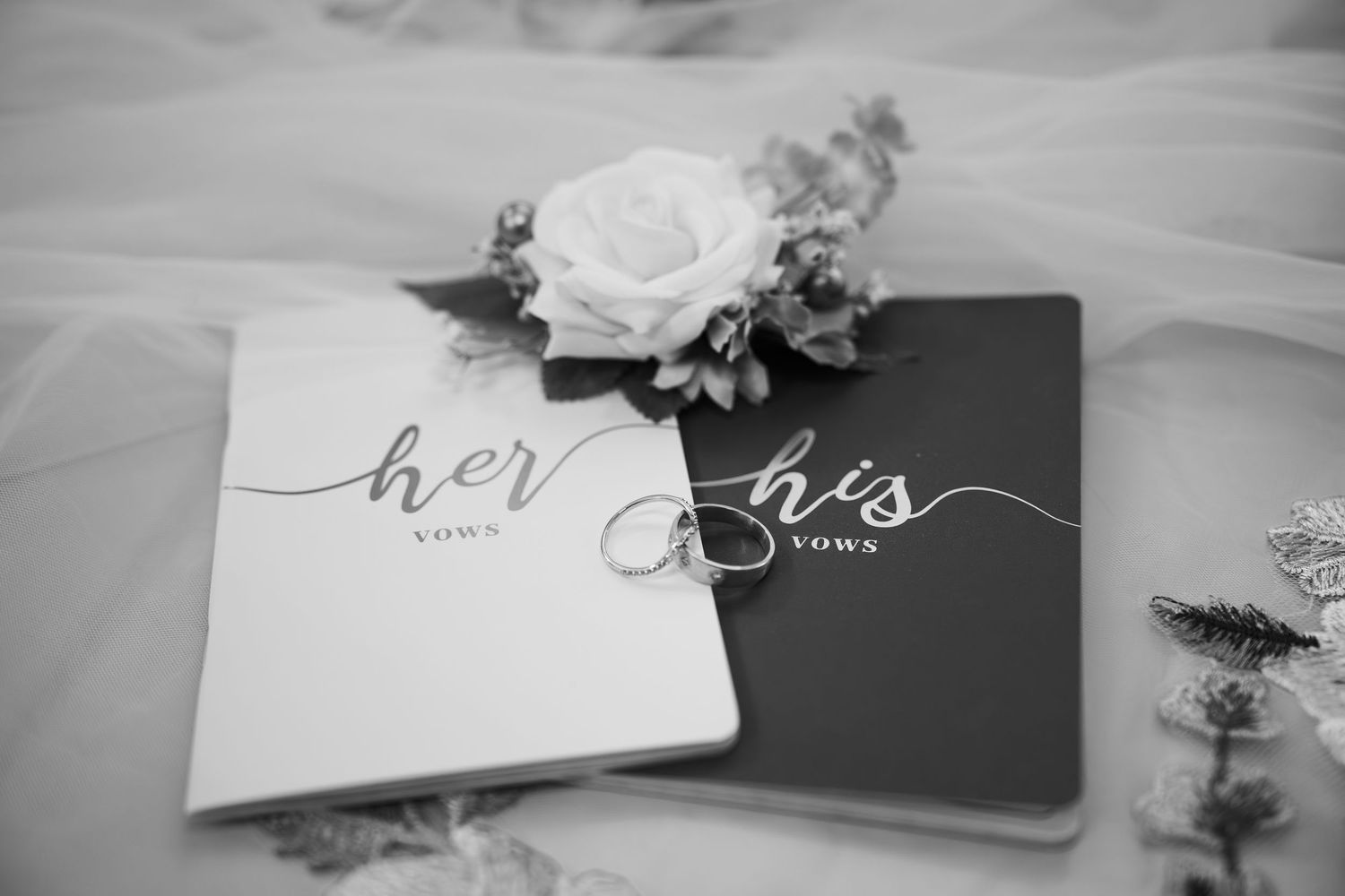 Black and white photo of her and his vow books with a white rose and wedding rings on a bridal veil.
