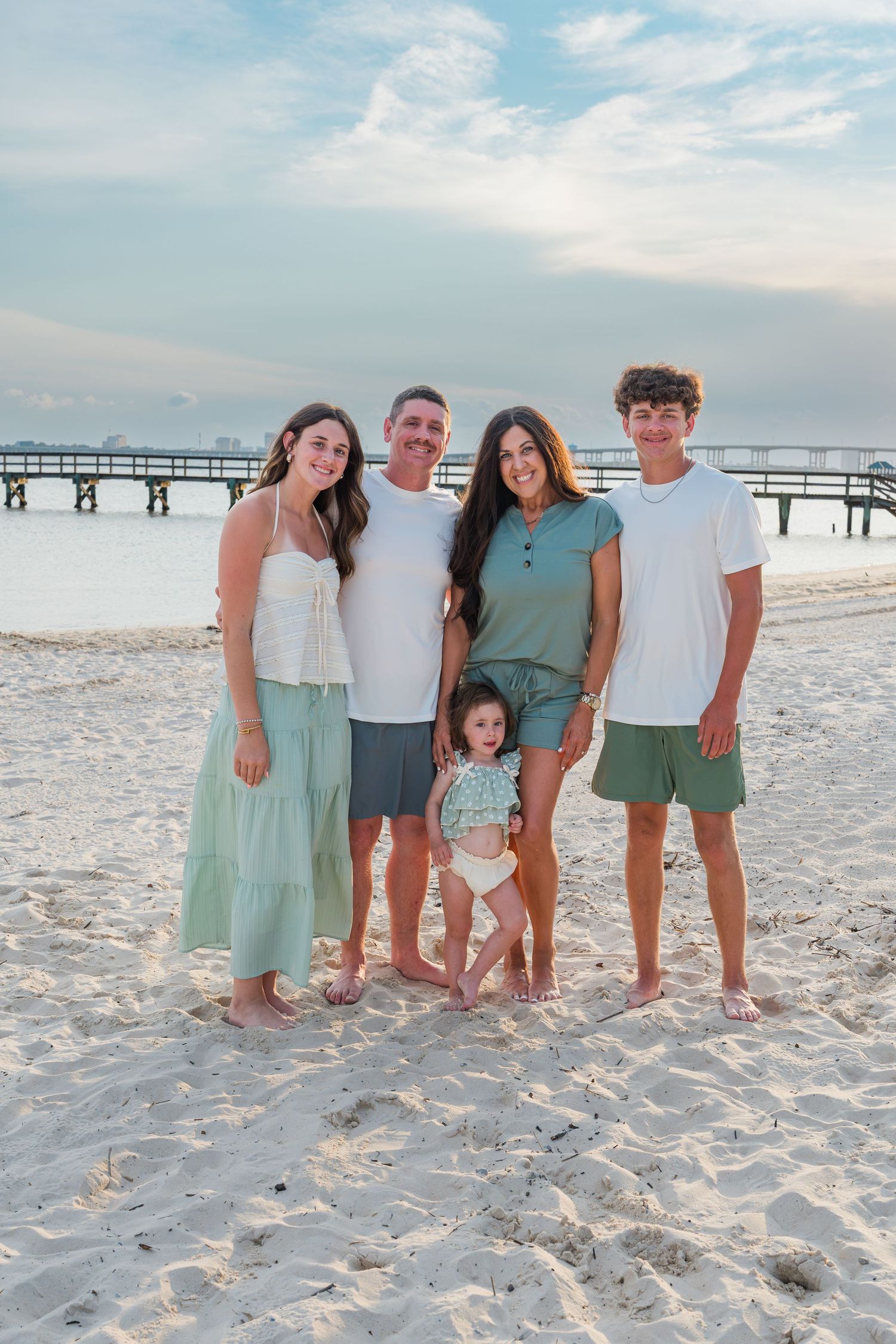 Family of five posing on a sandy beach at sunset, wearing coordinated white and green outfits, with a pier visible in the background.