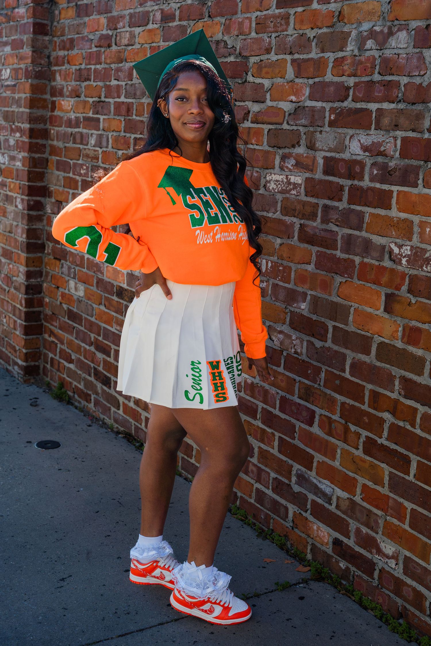 Graduate in orange university attire poses against brick wall wearing matching sneakers.