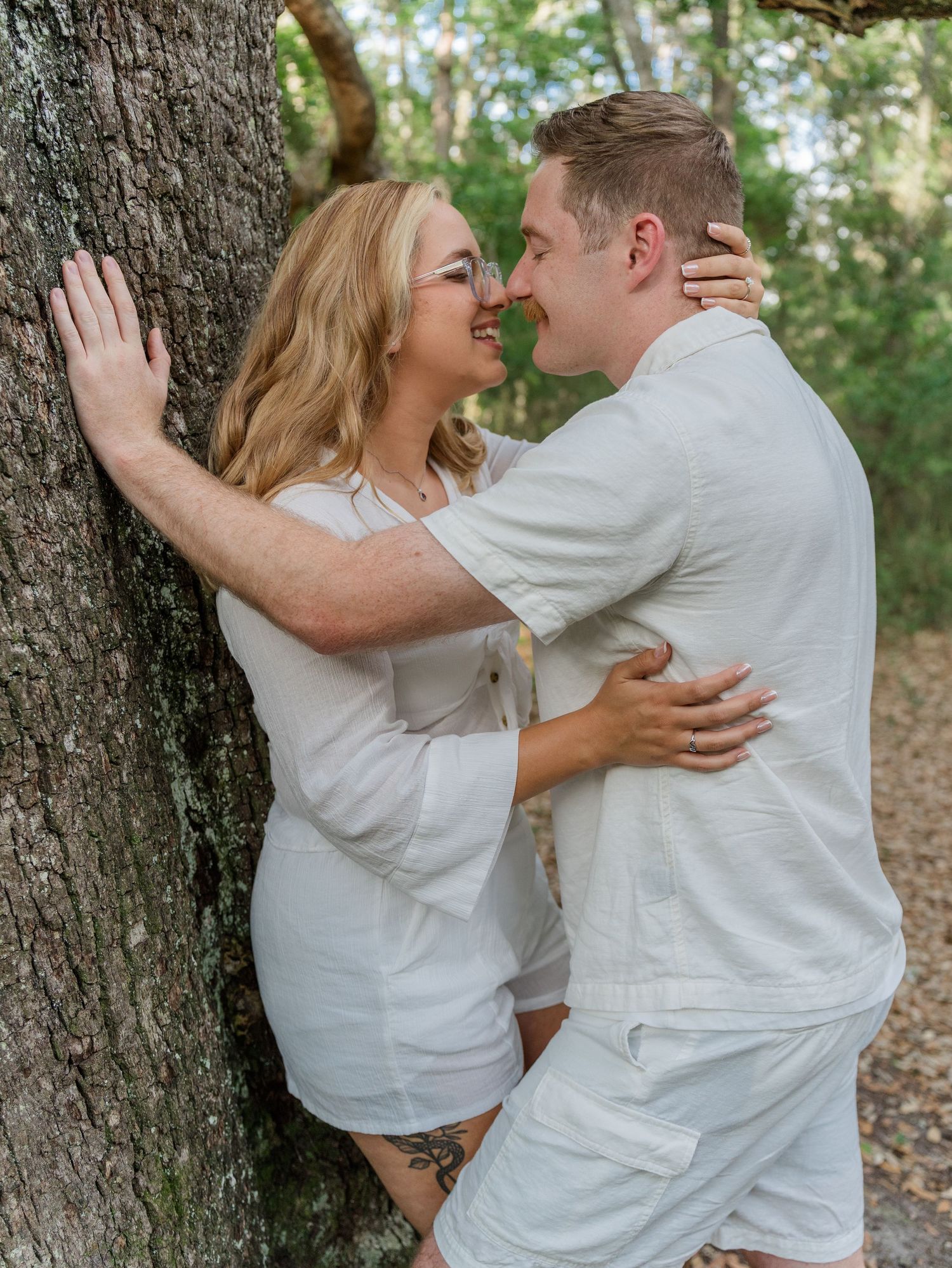 A couple embraces in white outfits against a tree backdrop during their engagement photoshoot.