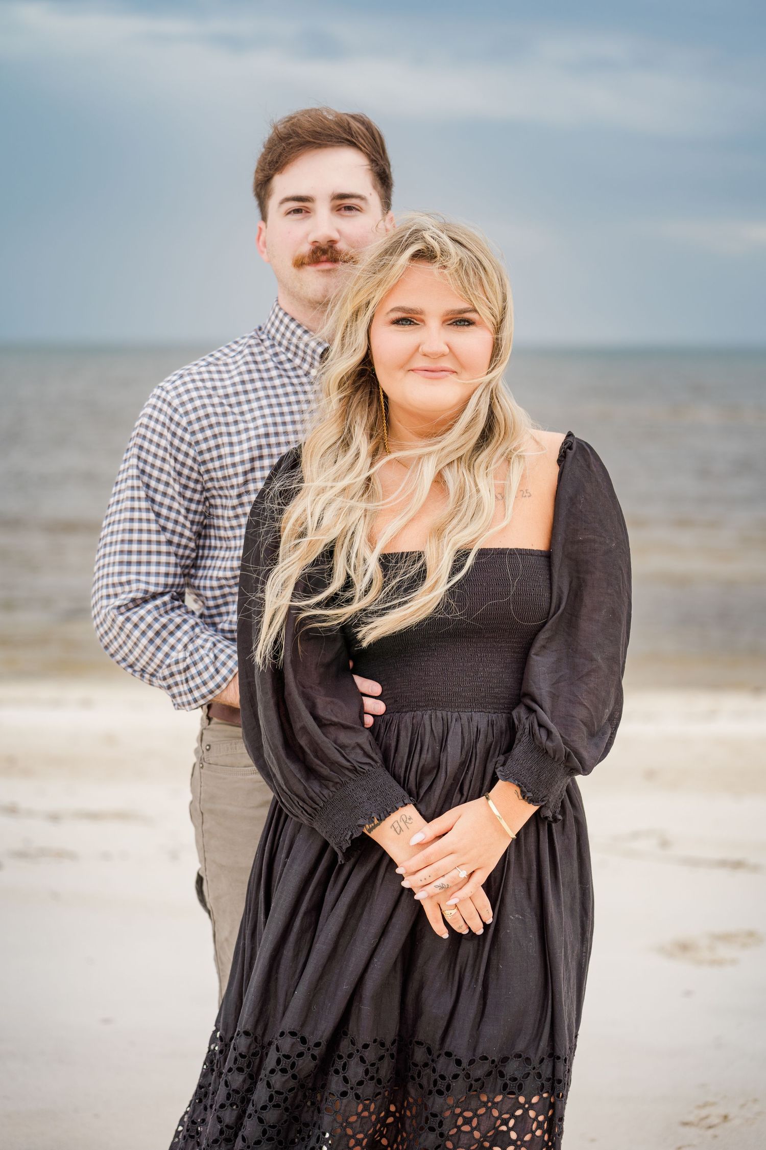 A couple embraces on a cloudy beach, with the woman wearing a flowing black dress.