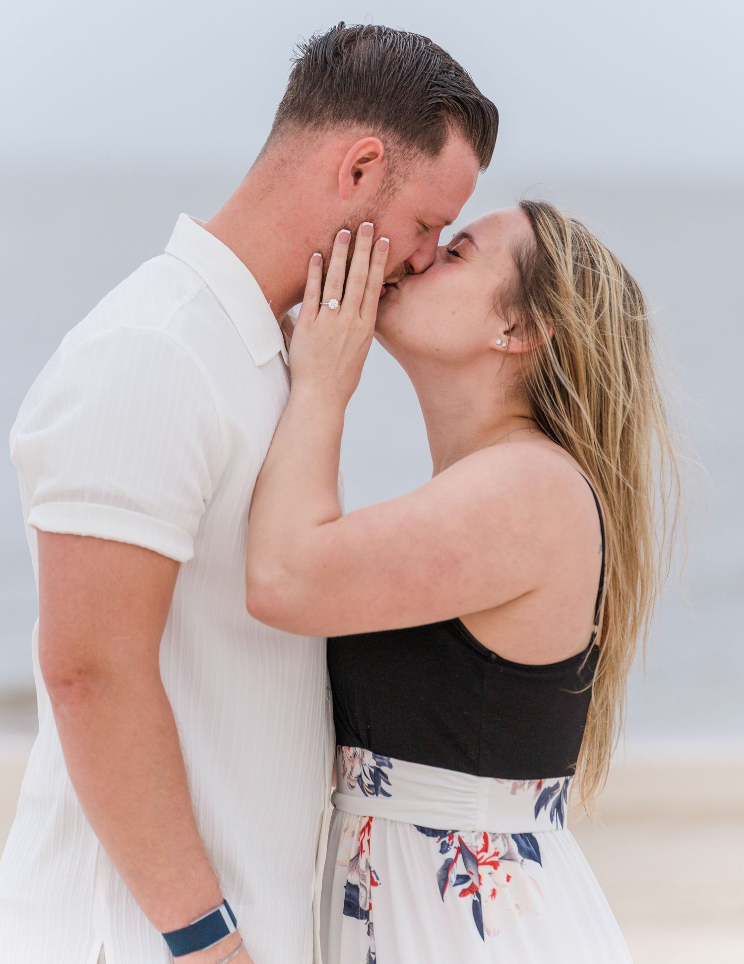 A newly engaged couple shares romantic kisses on the beach in matching white outfits.
