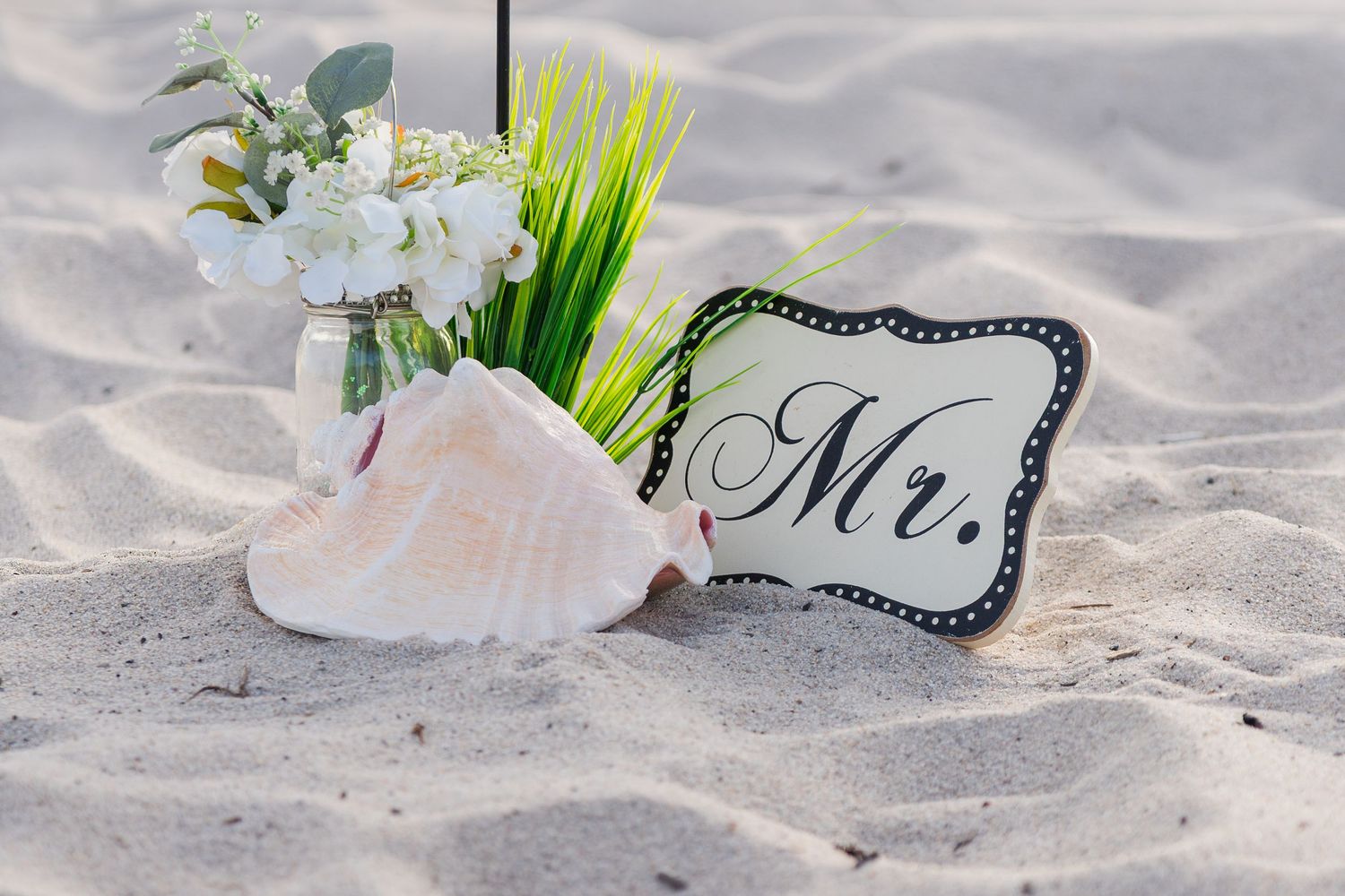 Beach wedding decor with a Mr. sign, seashell, and white floral arrangement displayed in white sand.