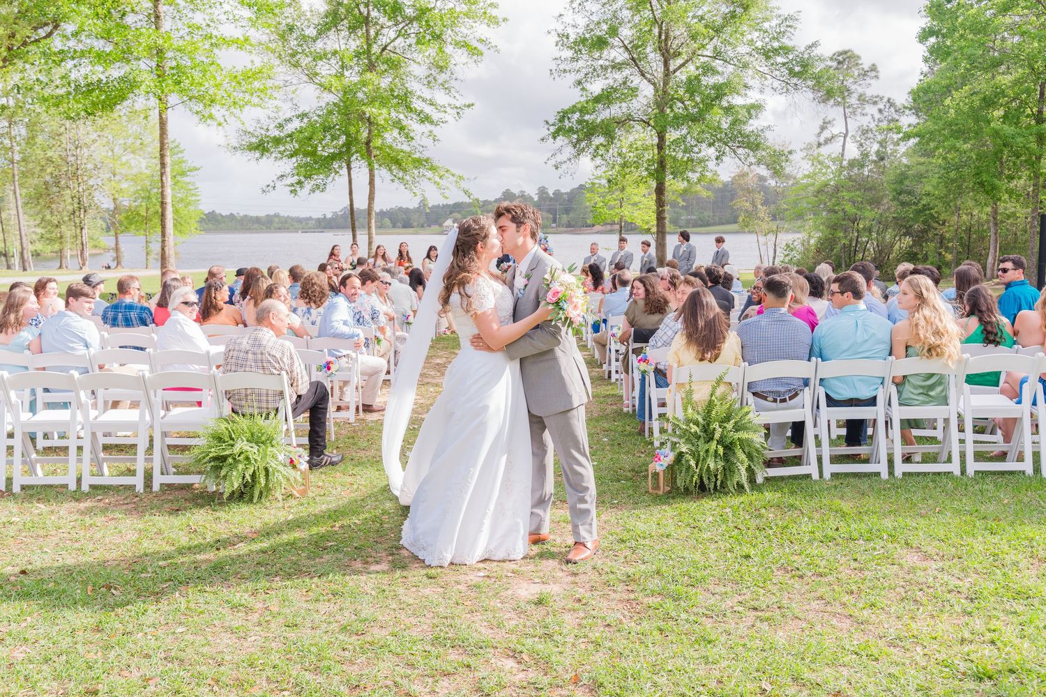 Bride and groom share first kiss at outdoor lakeside wedding ceremony with guests seated in white chairs.