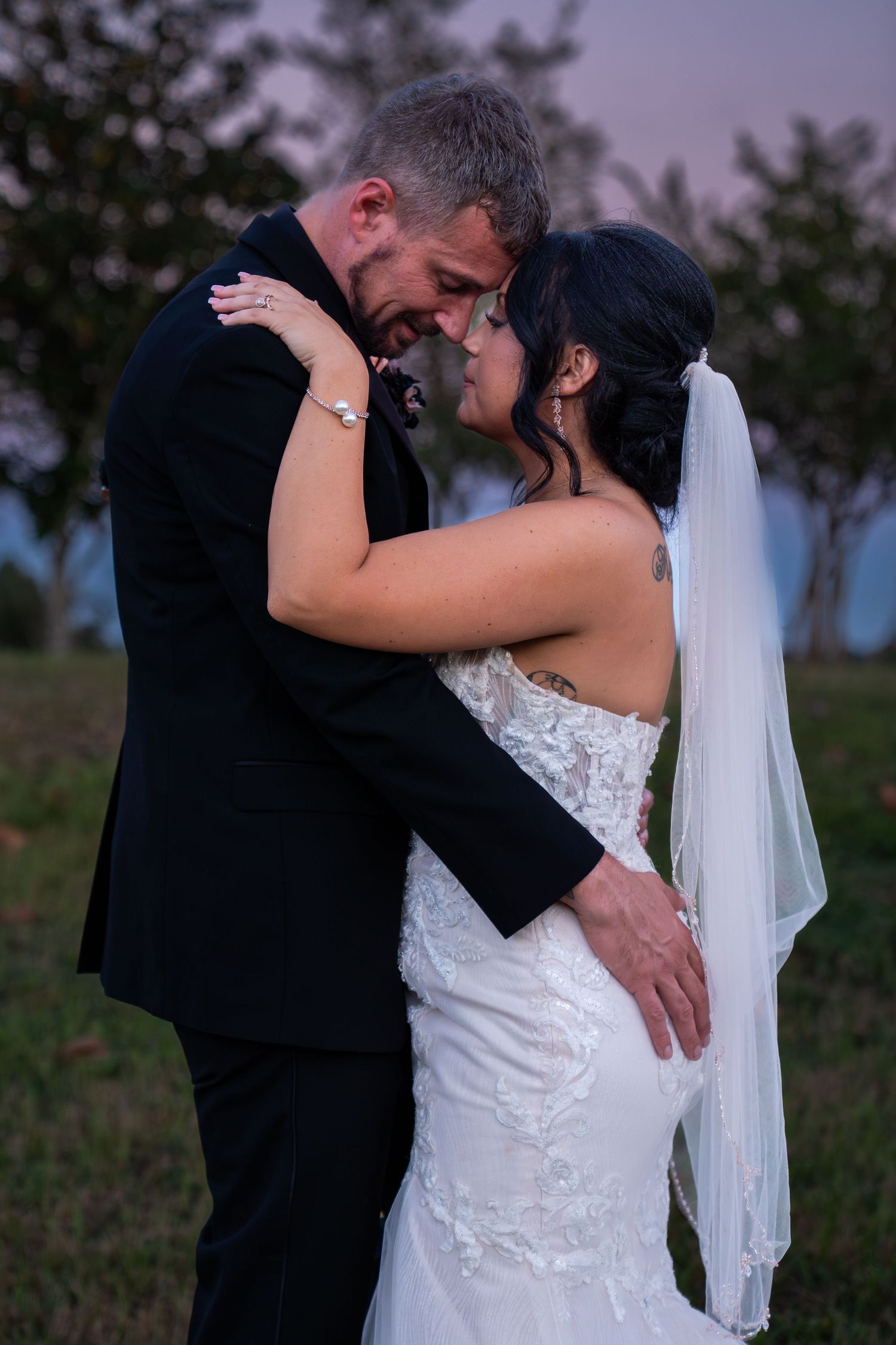 A romantic wedding couple embraces in a field at dusk, with the bride wearing a white lace gown and flowing veil.