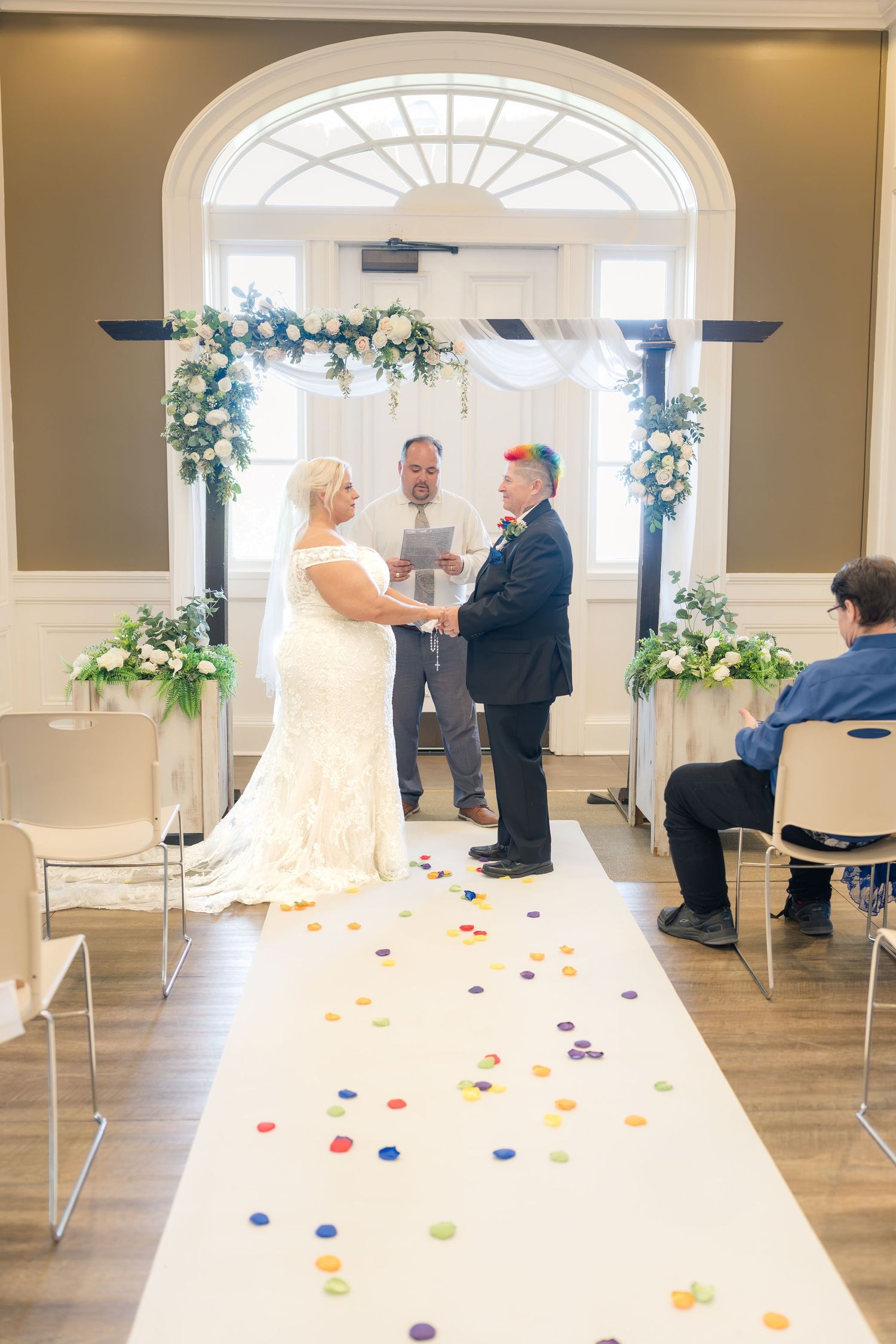 Bride and groom exchange vows at indoor wedding ceremony with floral arch, white aisle runner, and colorful confetti dots.