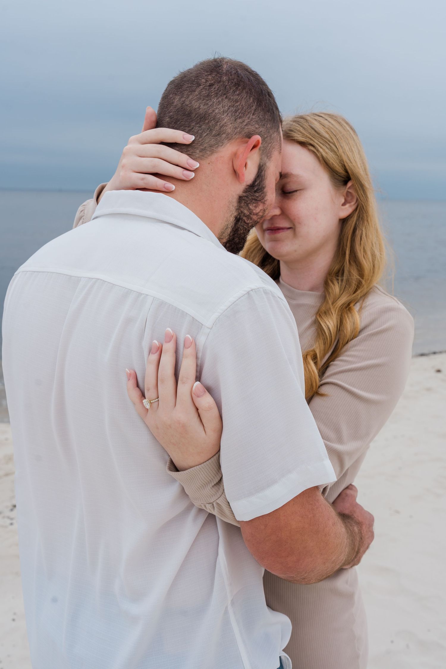 A romantic couple embraces tenderly on a beach with an overcast sky in the background.