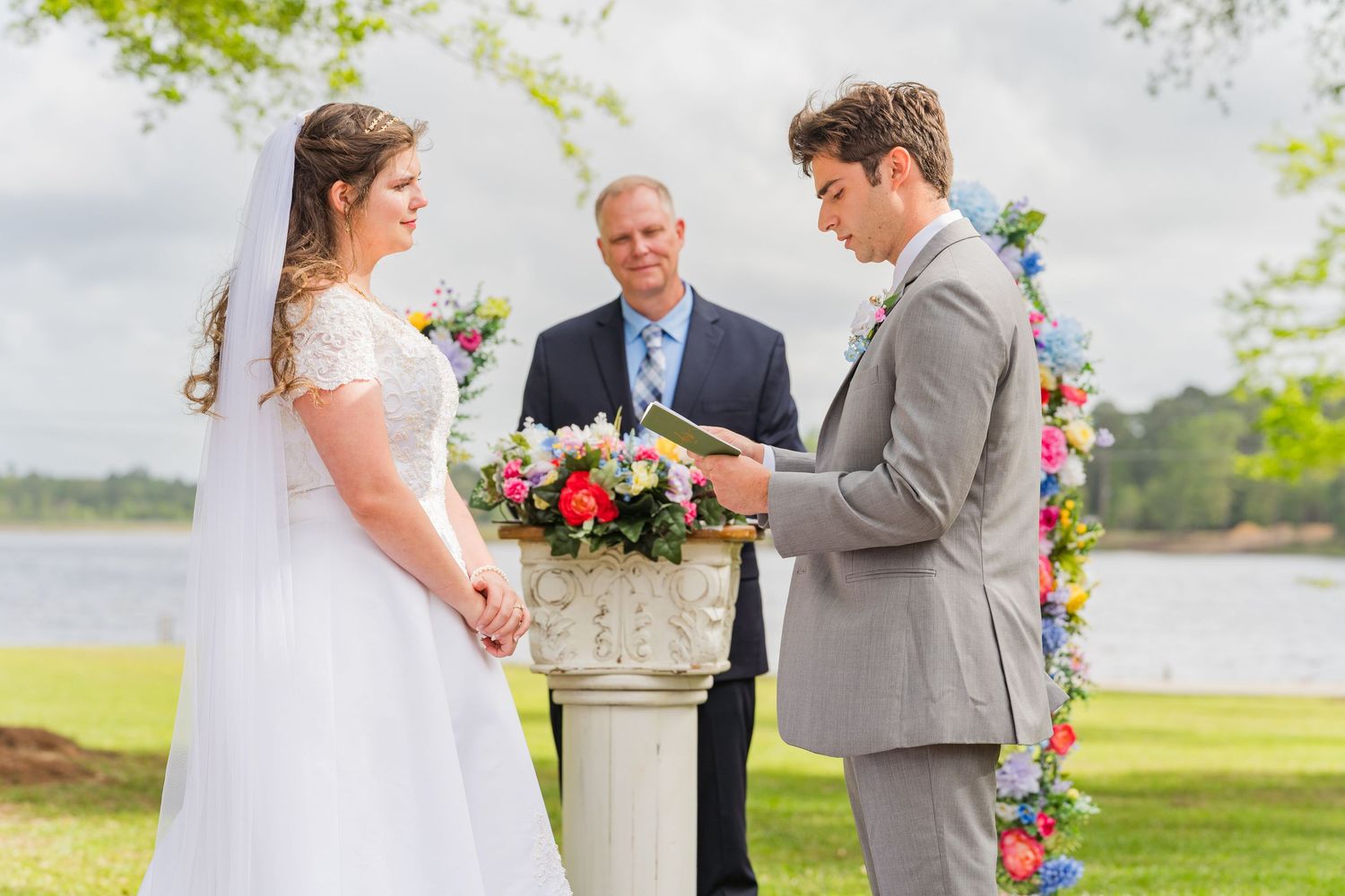Groom reads vows to bride during outdoor lakeside wedding ceremony with officiant and colorful floral arch.