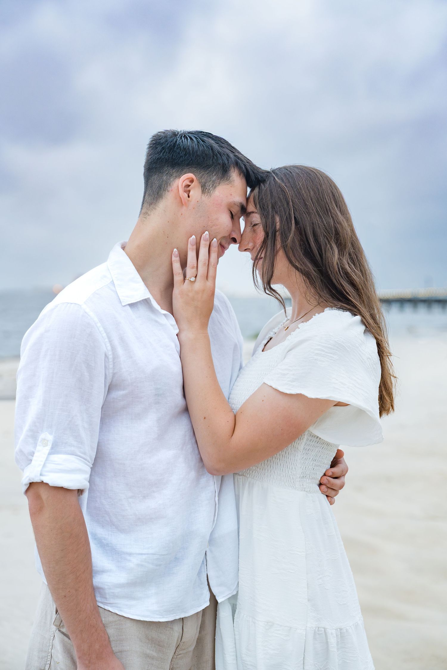 Romantic couple in white outfits sharing an intimate moment on a beach, touching foreheads tenderly.