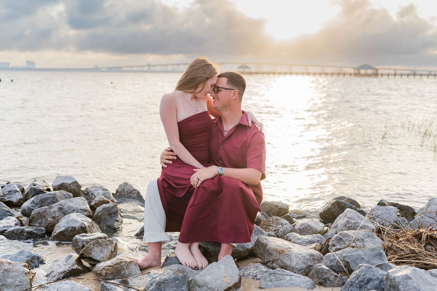 Couple sharing a romantic moment on rocky shoreline at sunset, woman in burgundy dress sitting on man's lap by the water.