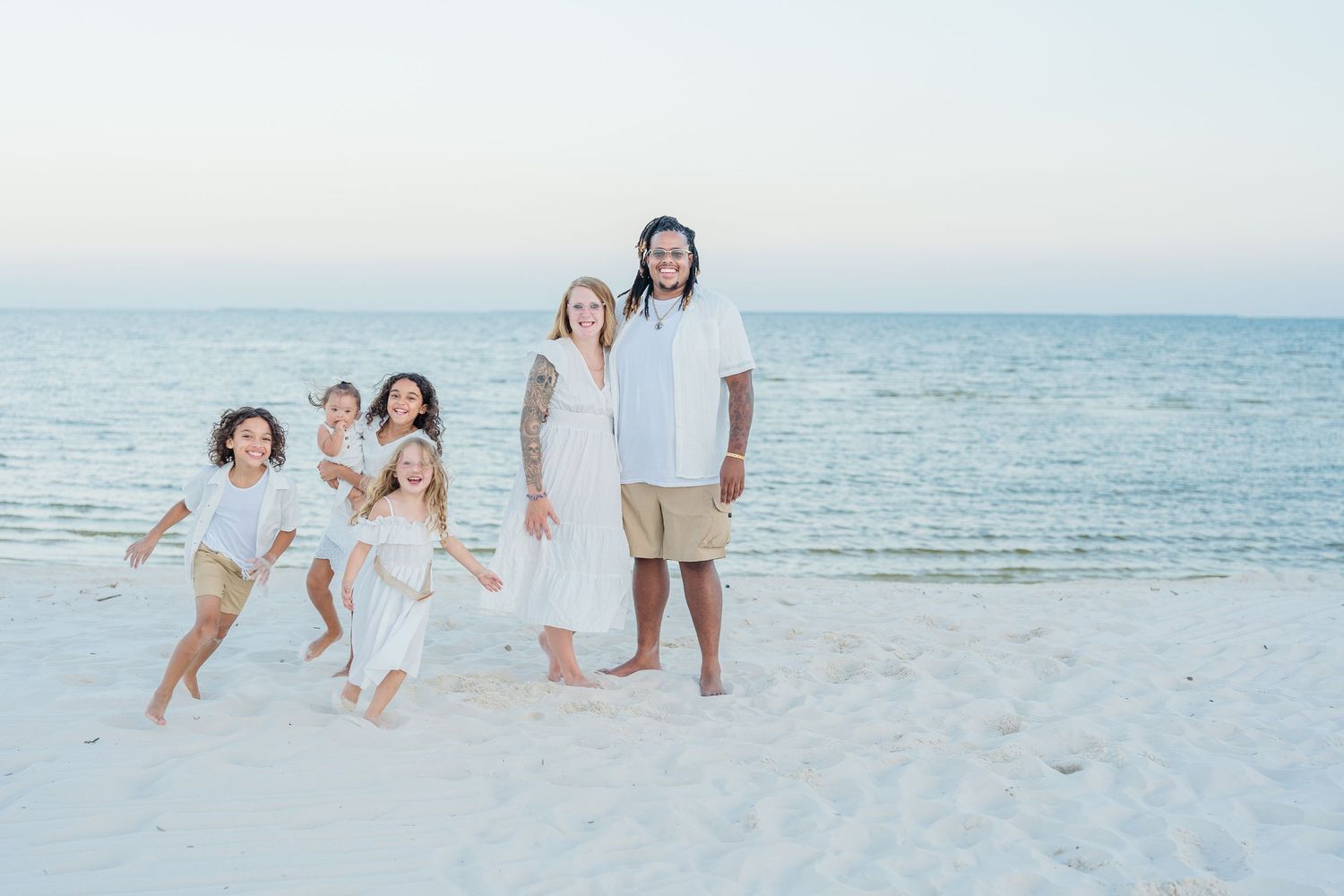 Joyful family in white outfits playing and posing on beach at sunset.